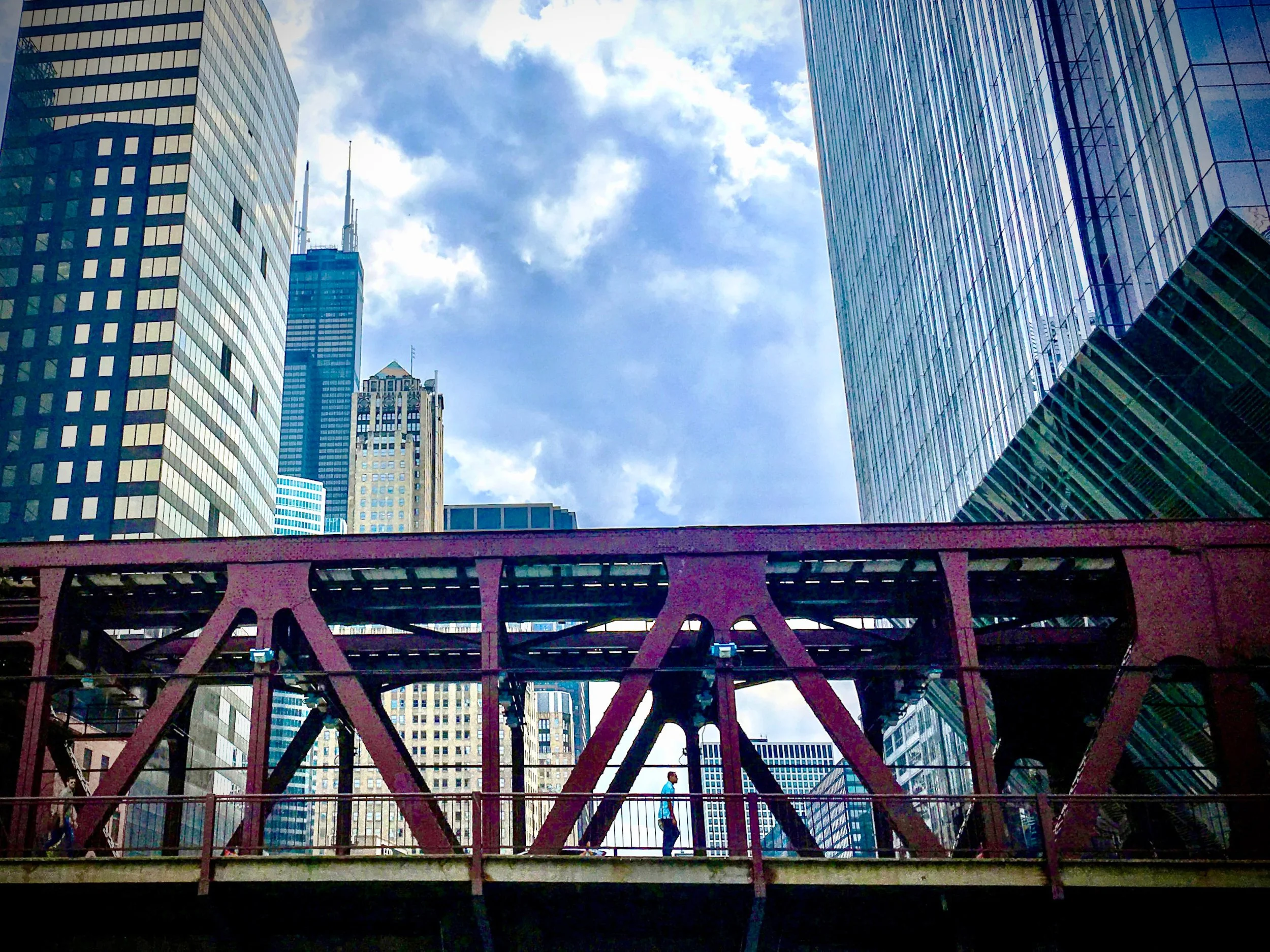 View of a man crossing a bridge with the Chicago skyline with tall glass skyscrapers under a partly cloudy blue sky behind him.