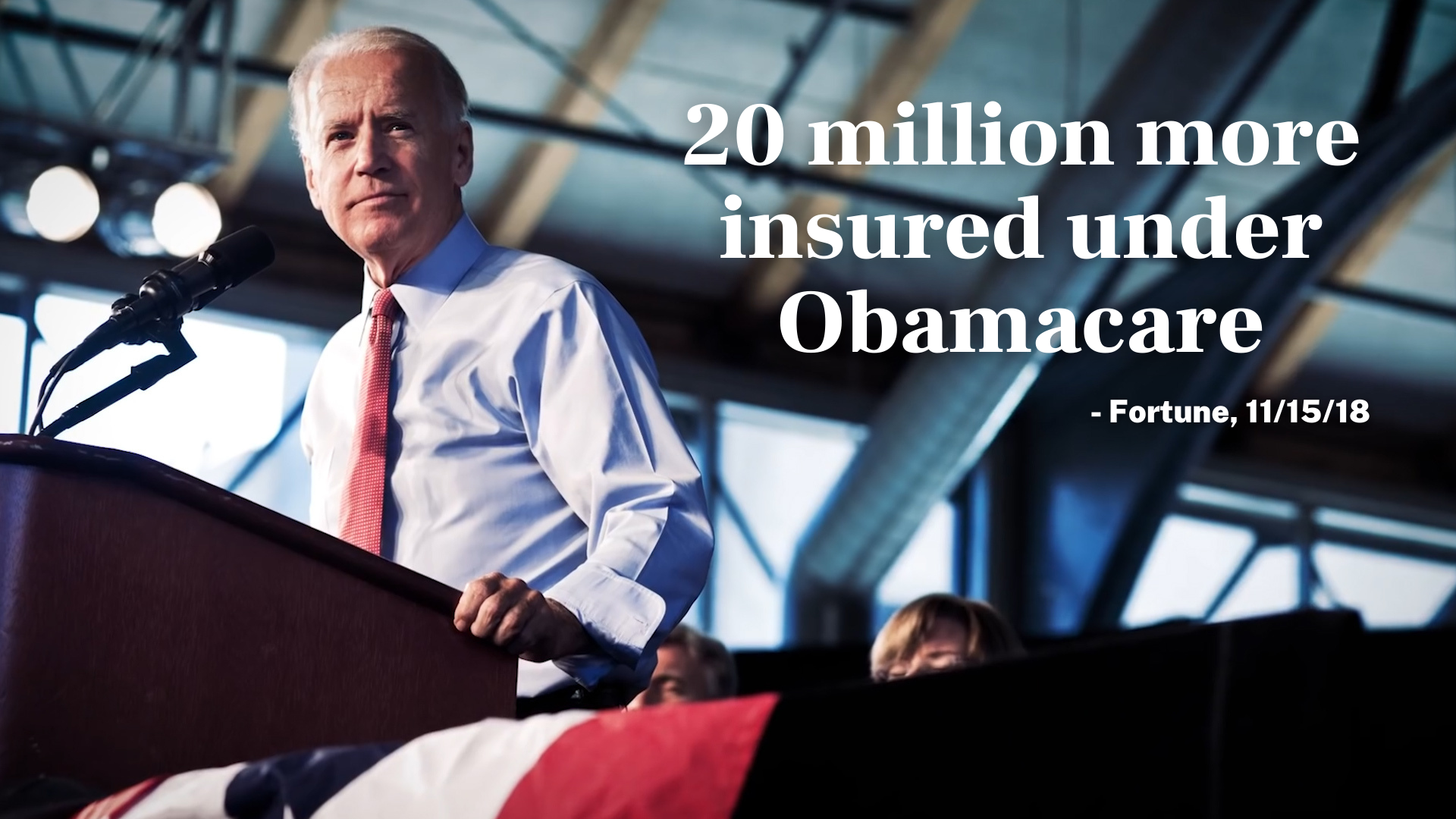 A still frame from an ad for President Joe Biden at a podium with a microphone, standing in an indoor setting with a modern industrial ceiling. Overlaid text reads: '20 million more insured under Obamacare - Fortune, 11/15/18.'