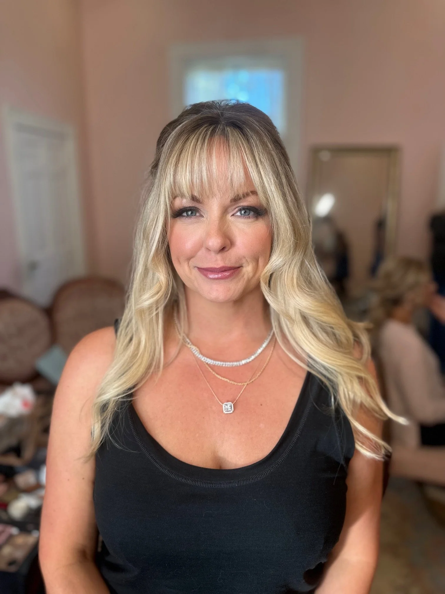 A woman with blonde hair styled in loose waves and bangs, wearing a black sleeveless top and layered silver necklaces, smiling in a room with pink-colored walls and blurred background.