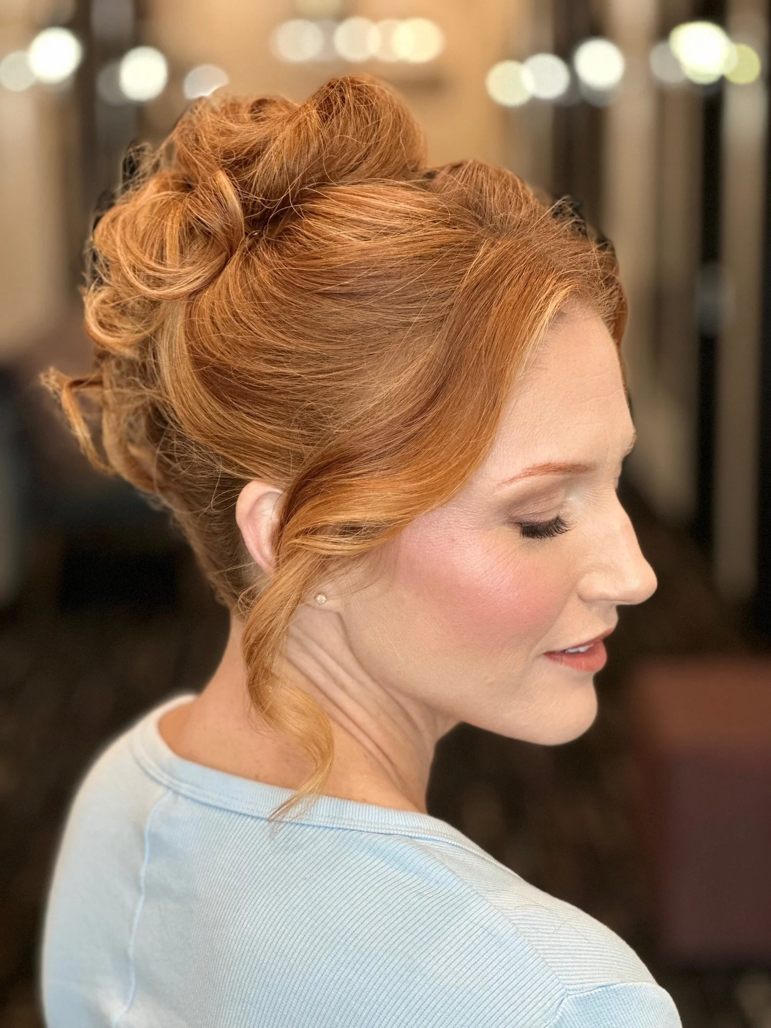 A woman with red hair styled in an elegant updo, with soft curls and loose strands, wearing a light blue top, with her eyes closed and a slight smile.