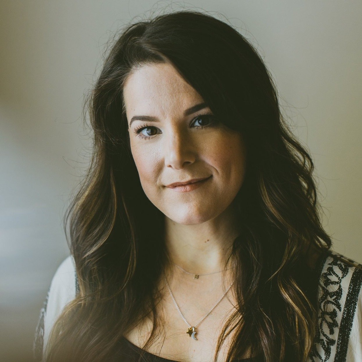 Portrait of a woman with long wavy brown hair, fair skin, blue eyes, and subtle makeup, wearing necklaces and a patterned black-and-white top, standing against a neutral background.