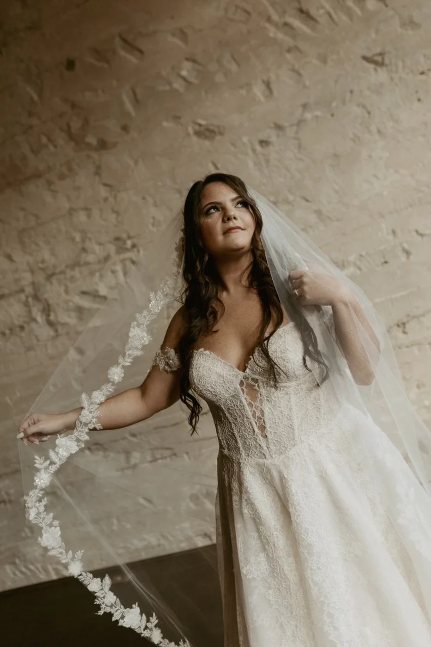 Bride in a lace wedding dress holding a veil with floral embroidery, standing against a textured brick wall.