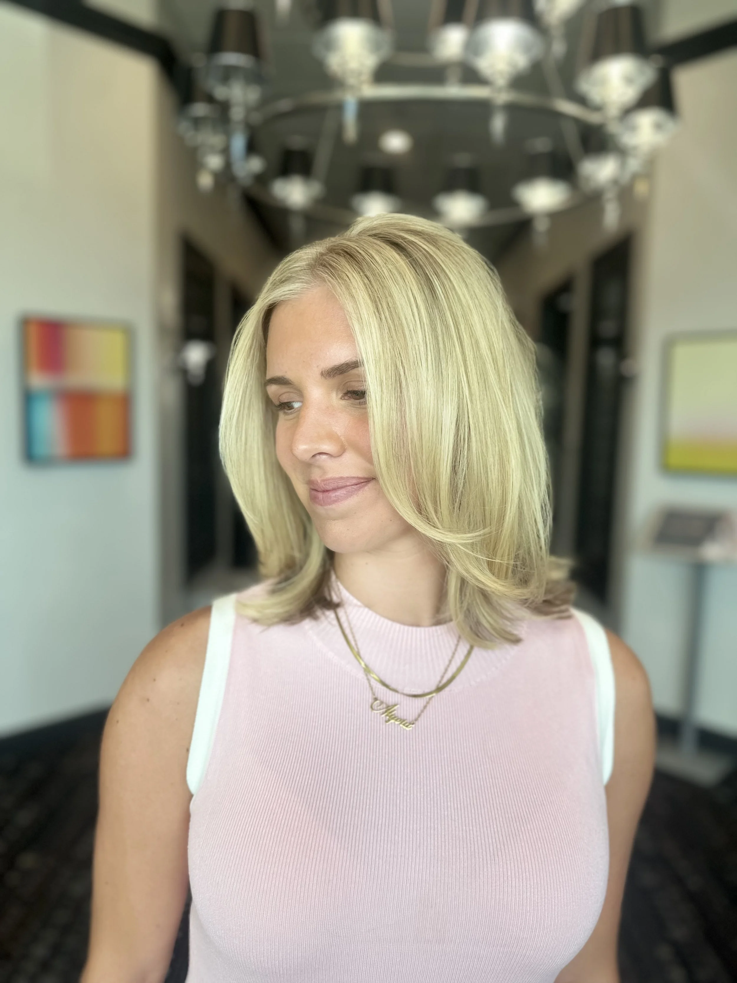A woman with blonde hair and light skin wearing a sleeveless pink top and gold necklace, standing in front of a blurred background of a hallway with a large chandelier.