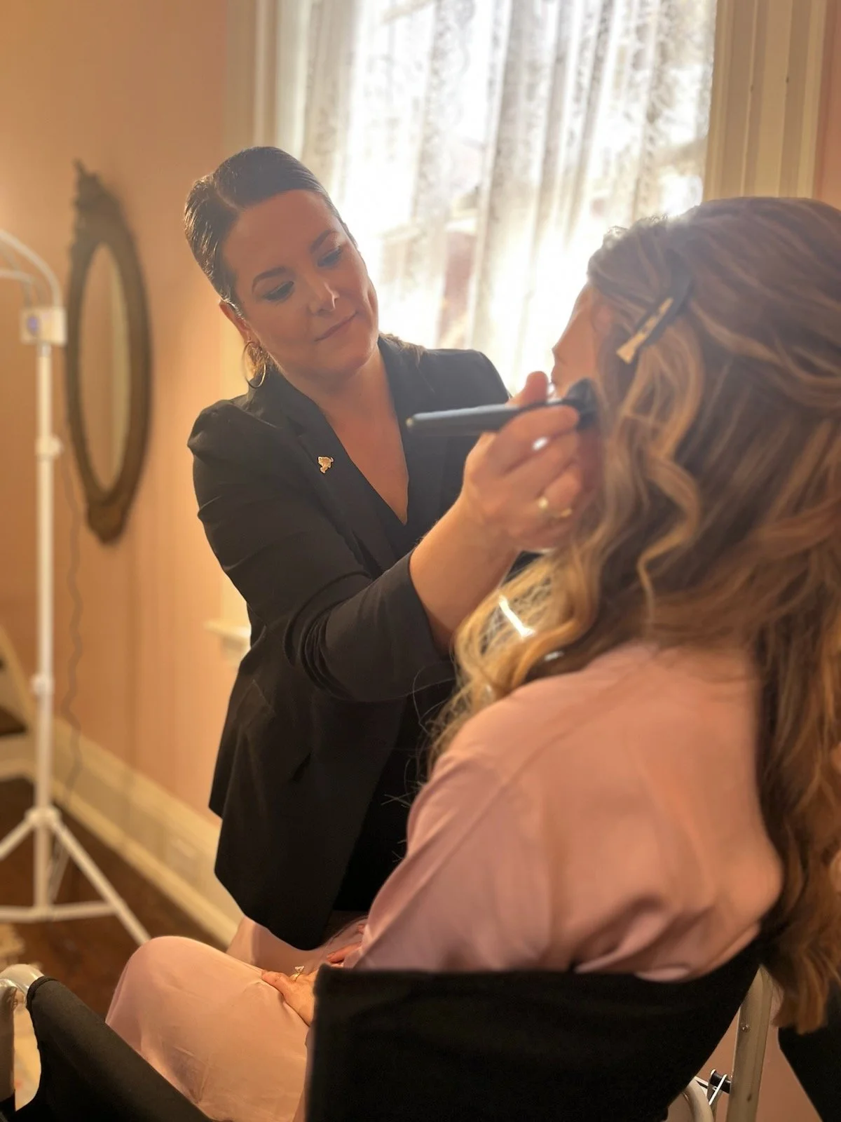 Makeup artist applying makeup to a woman seated in a chair, with sunlight coming through a window in the background.