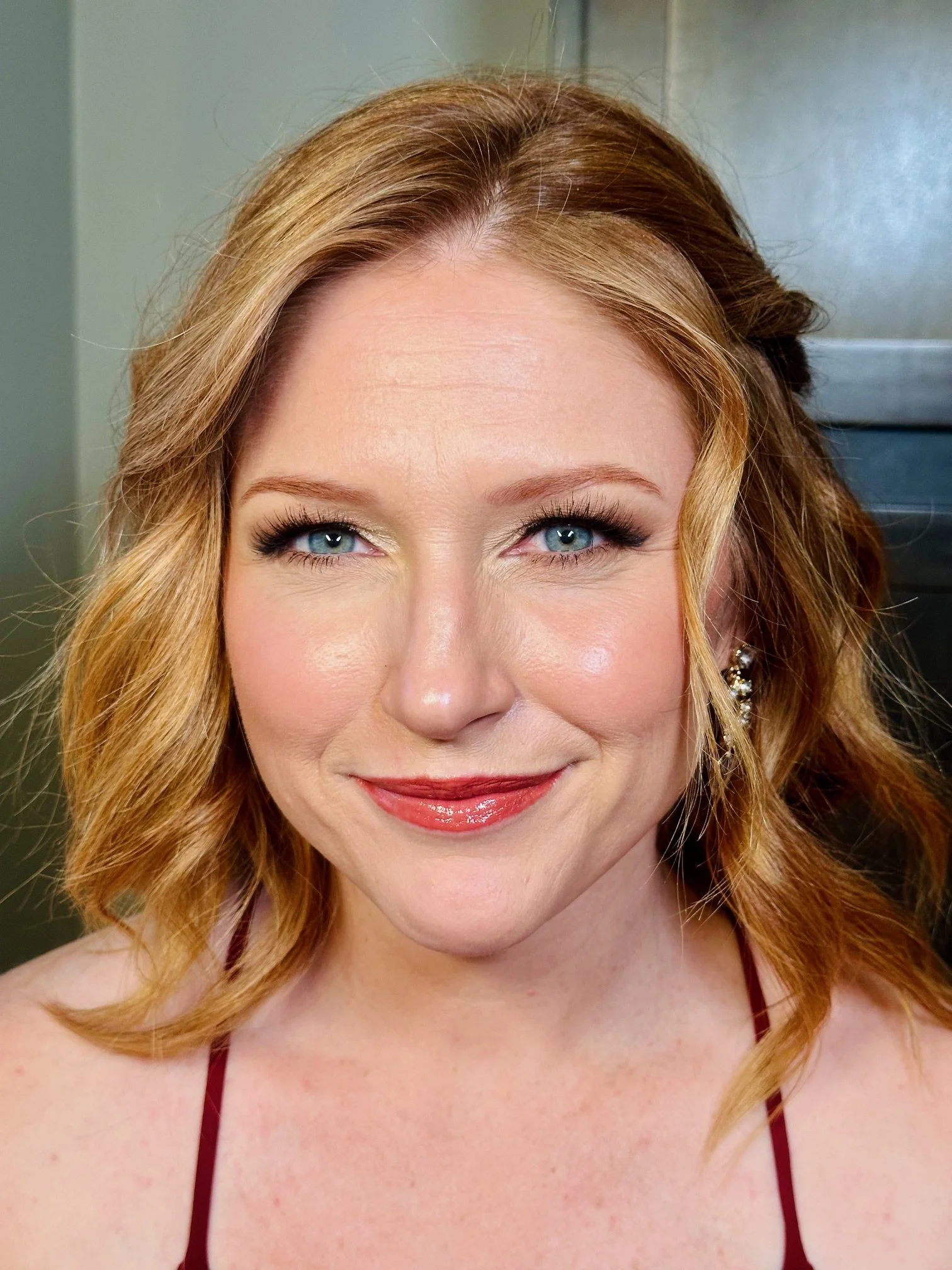 Close-up of a woman with red hair, blue eyes, and a smile, wearing makeup and earrings, with a neutral background.