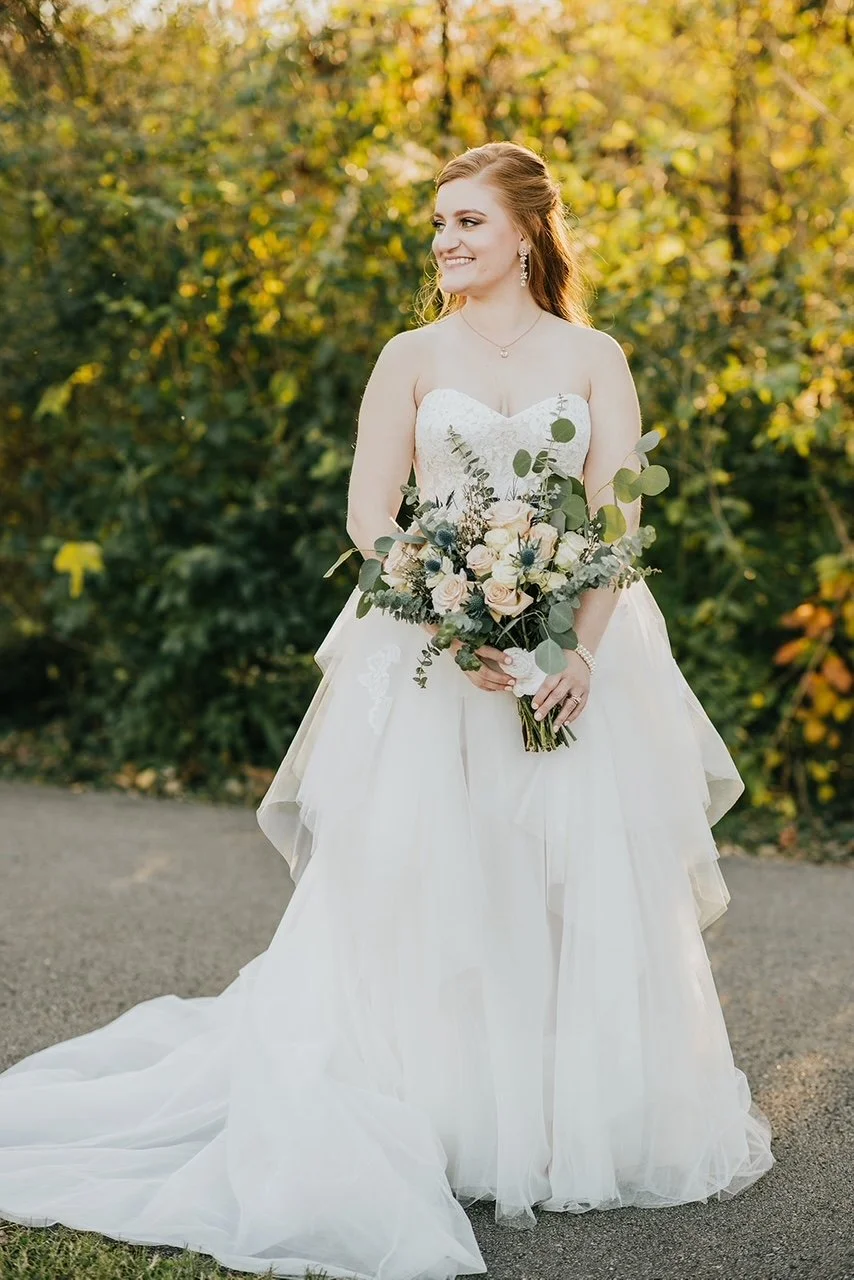 A bride in a strapless wedding gown holding a bouquet of white and blush roses with greenery, standing outdoors with a backdrop of autumn trees.