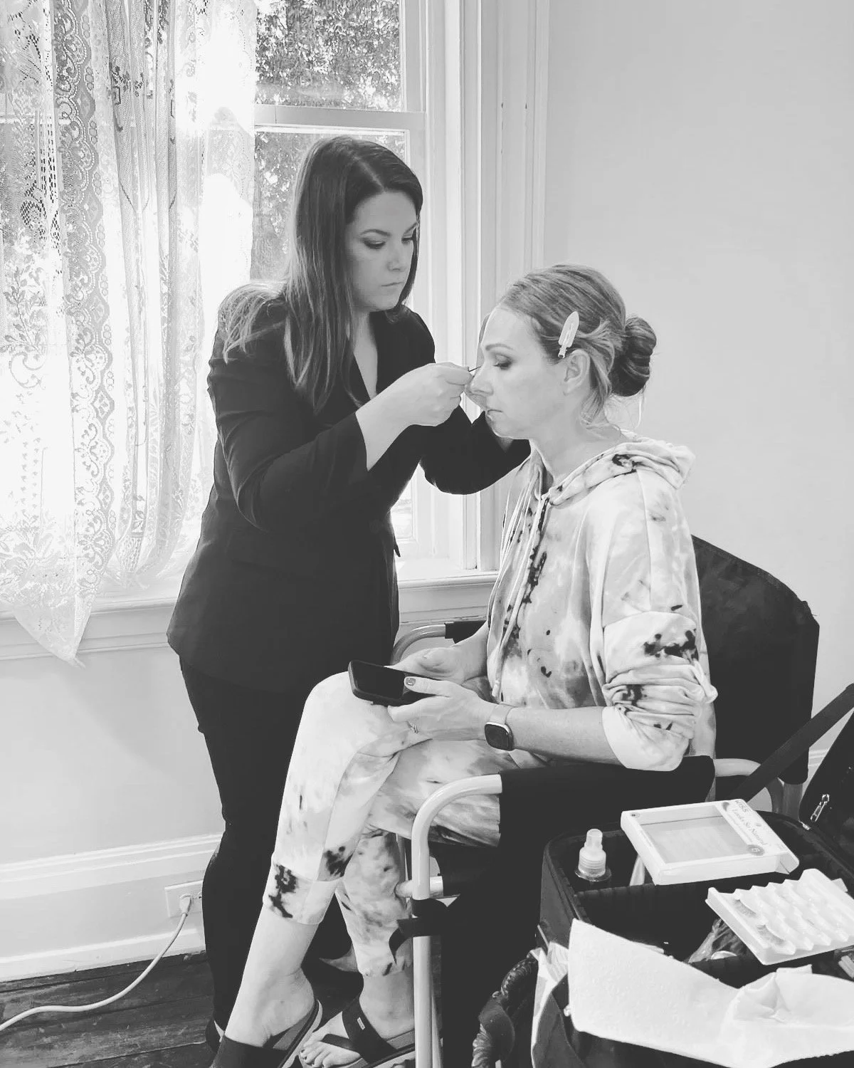 A woman sitting in a chair getting her makeup done by a makeup artist in a room with lace curtains and a window.