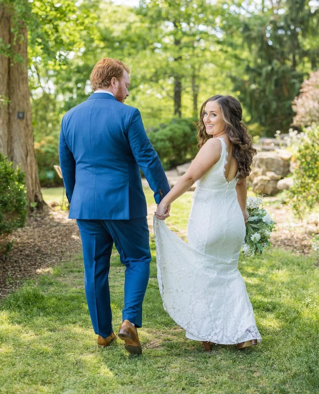 A bride and groom holding hands outdoors in a green garden, with the bride smiling at the camera and holding a bouquet.