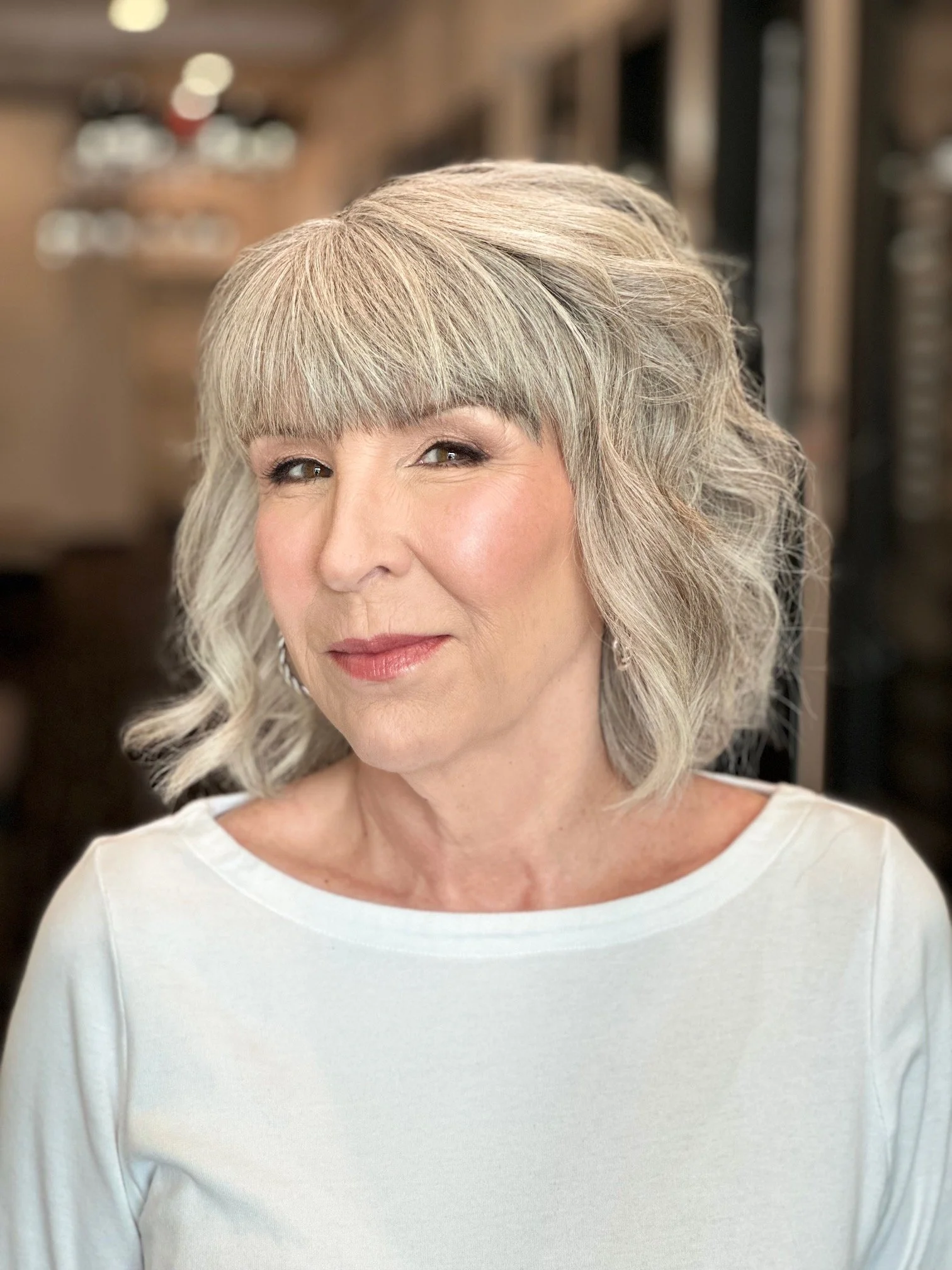 A woman with shoulder-length gray hair styled in loose waves and straight bangs, wearing a white top, smiling softly, with a blurred background of a hair salon.