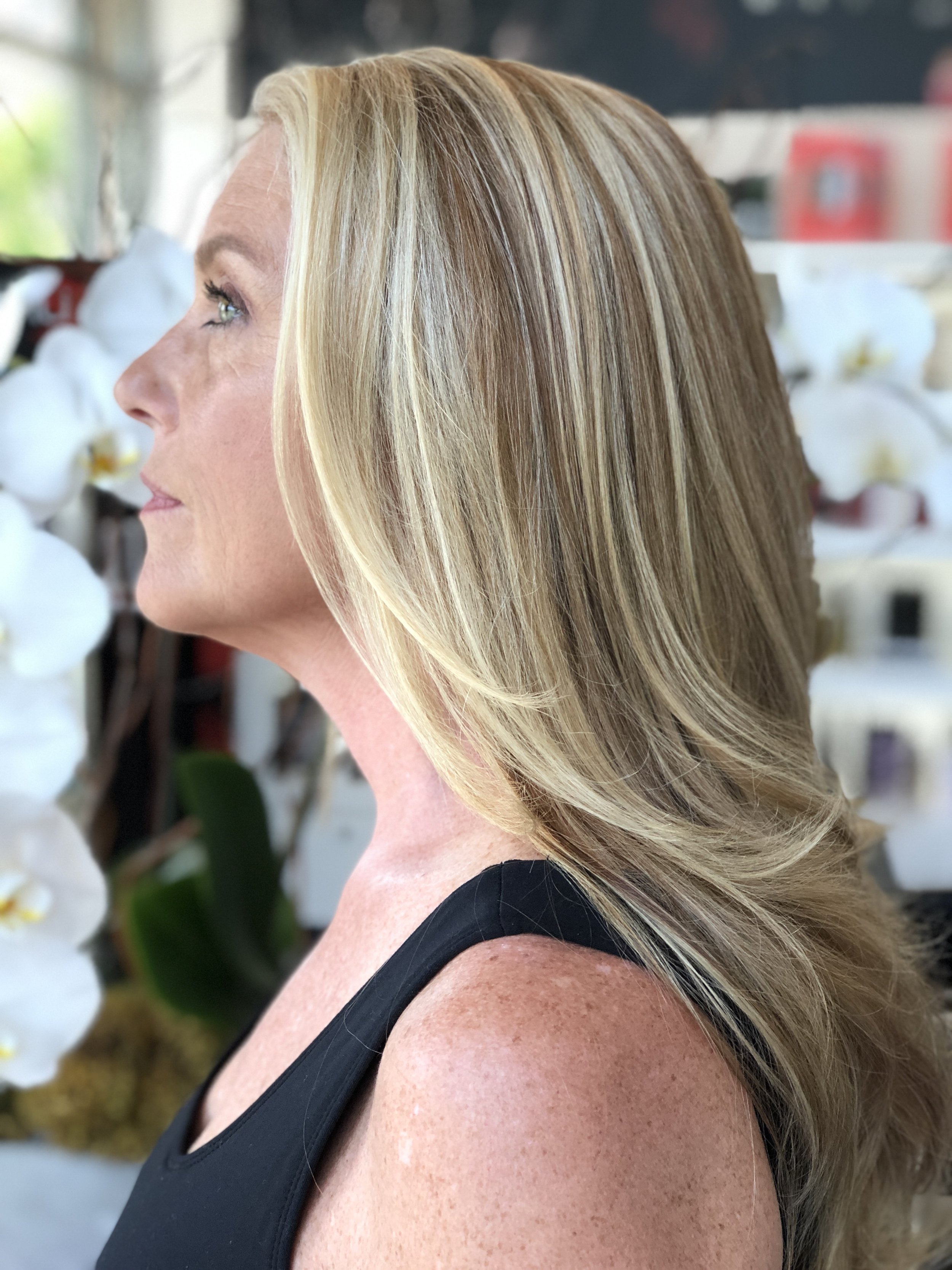 Close-up of a woman with long blonde hair wearing a black sleeveless top, with white orchids in the background.
