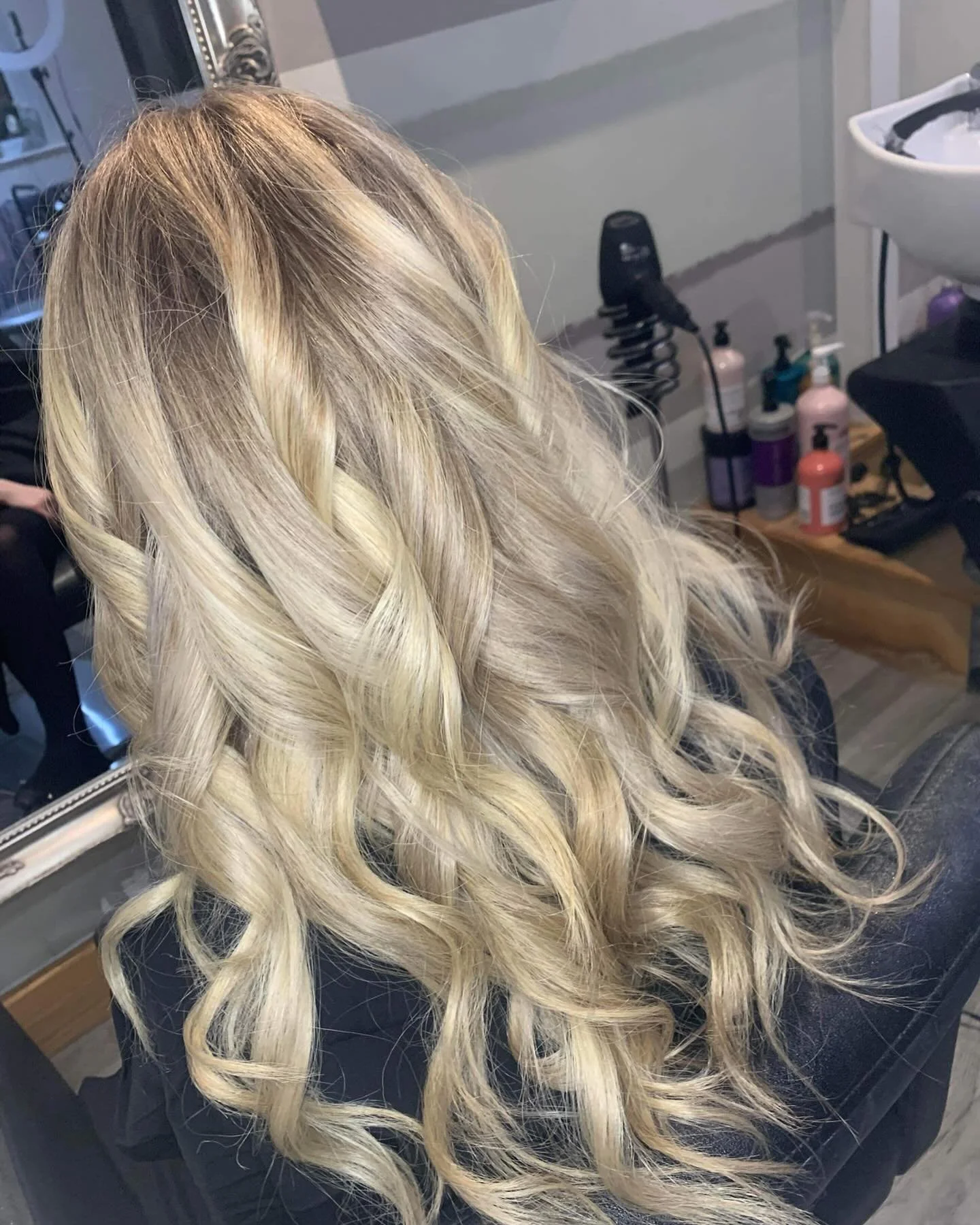 Blonde woman with long, curly hair sitting in a salon chair.