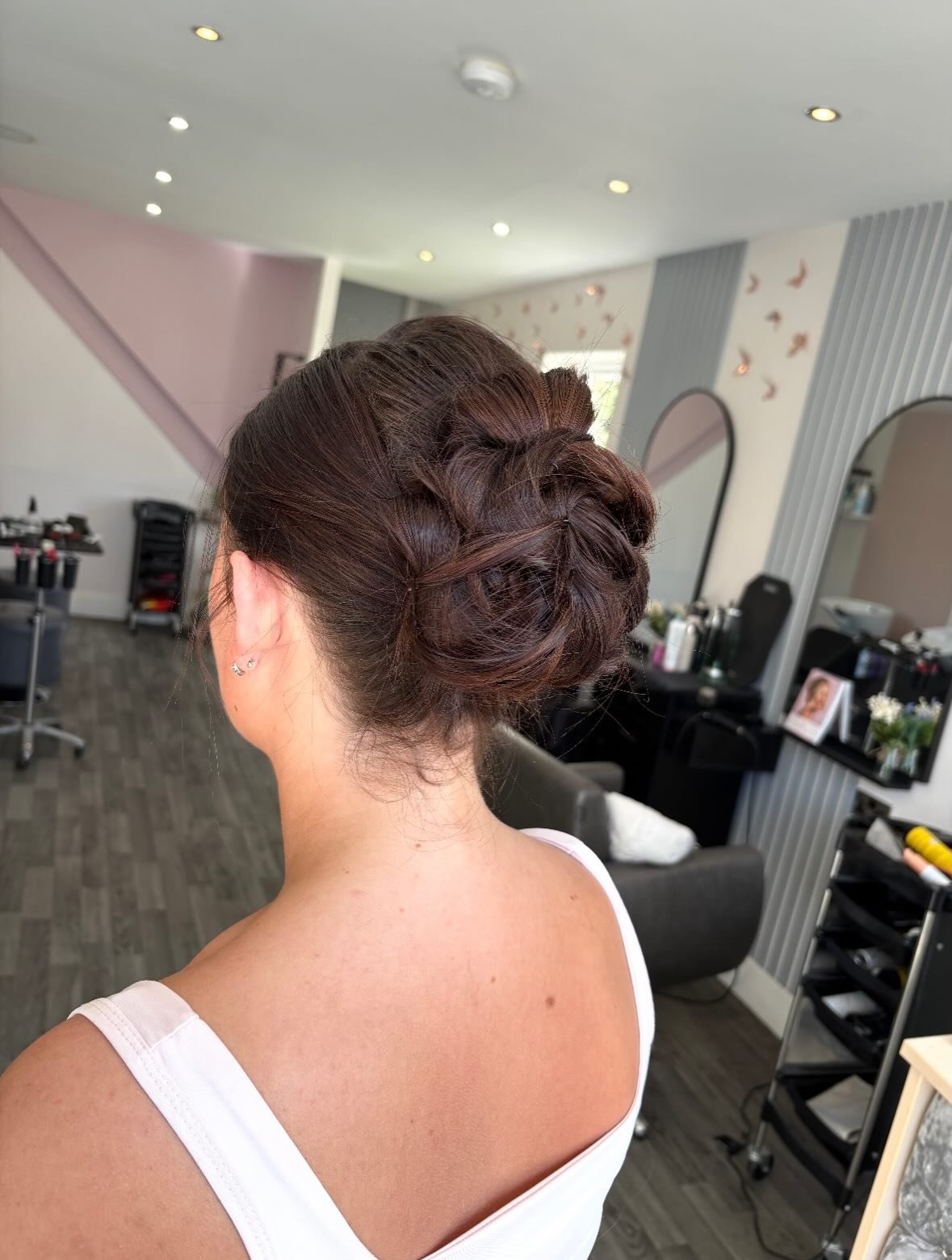 A woman with brunette hair styled in an elegant updo, sitting in a salon, with mirrors, salon equipment, and a hairstylist's station visible in the background.