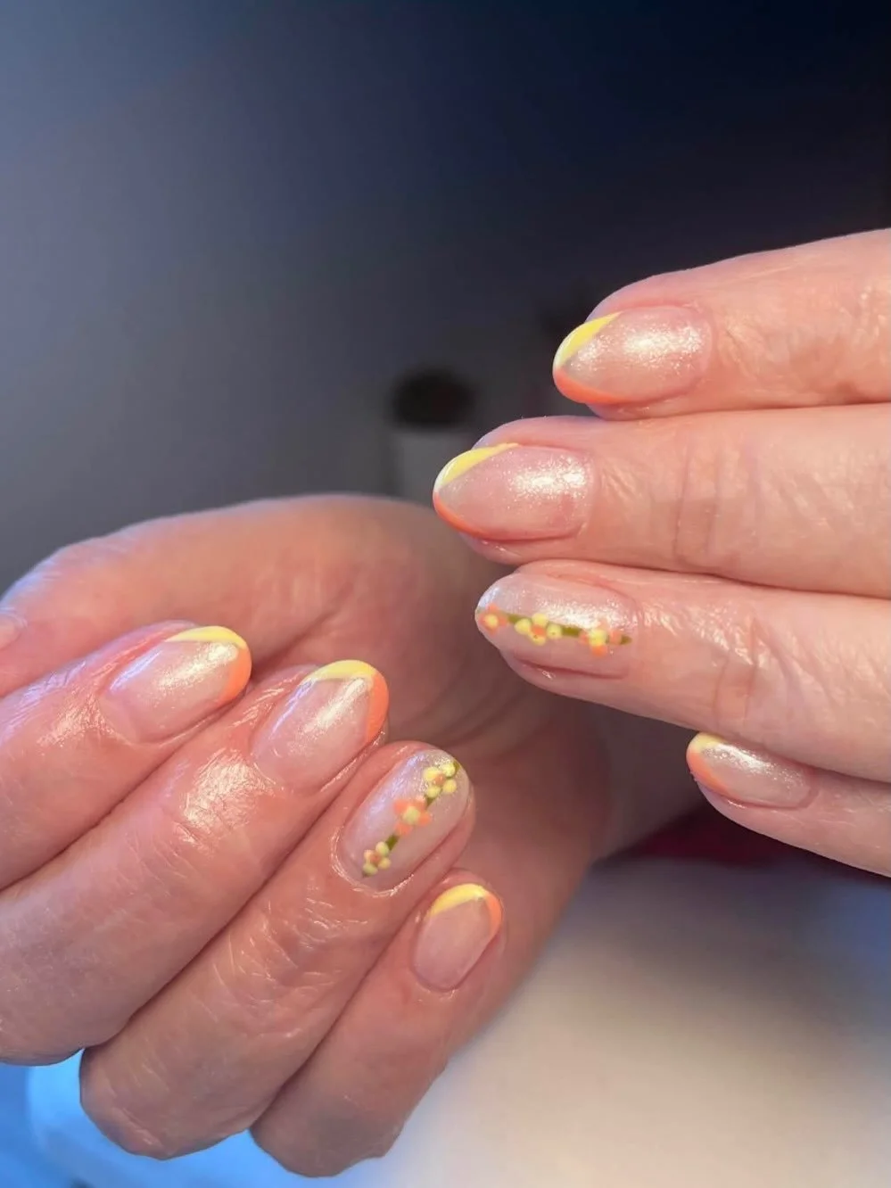 Close-up of hands with manicured nails featuring a clear base with yellow and orange French tips and small yellow and orange floral designs.