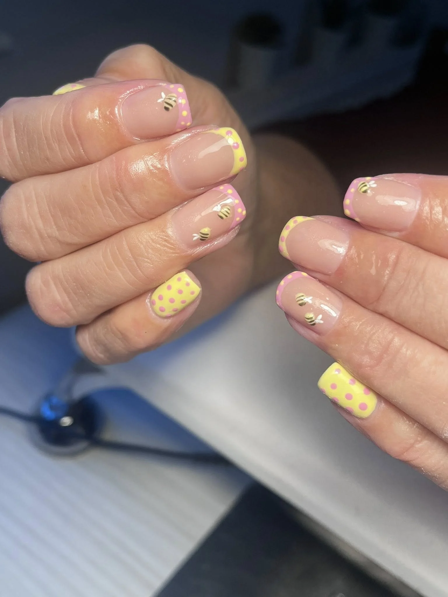 Close-up of hands with colorful, bee-themed nail art featuring yellow and pink polka dots and small bee designs. Award winning Lisa B Hair and Beauty Salon in Carlisle, Cumbria UK.