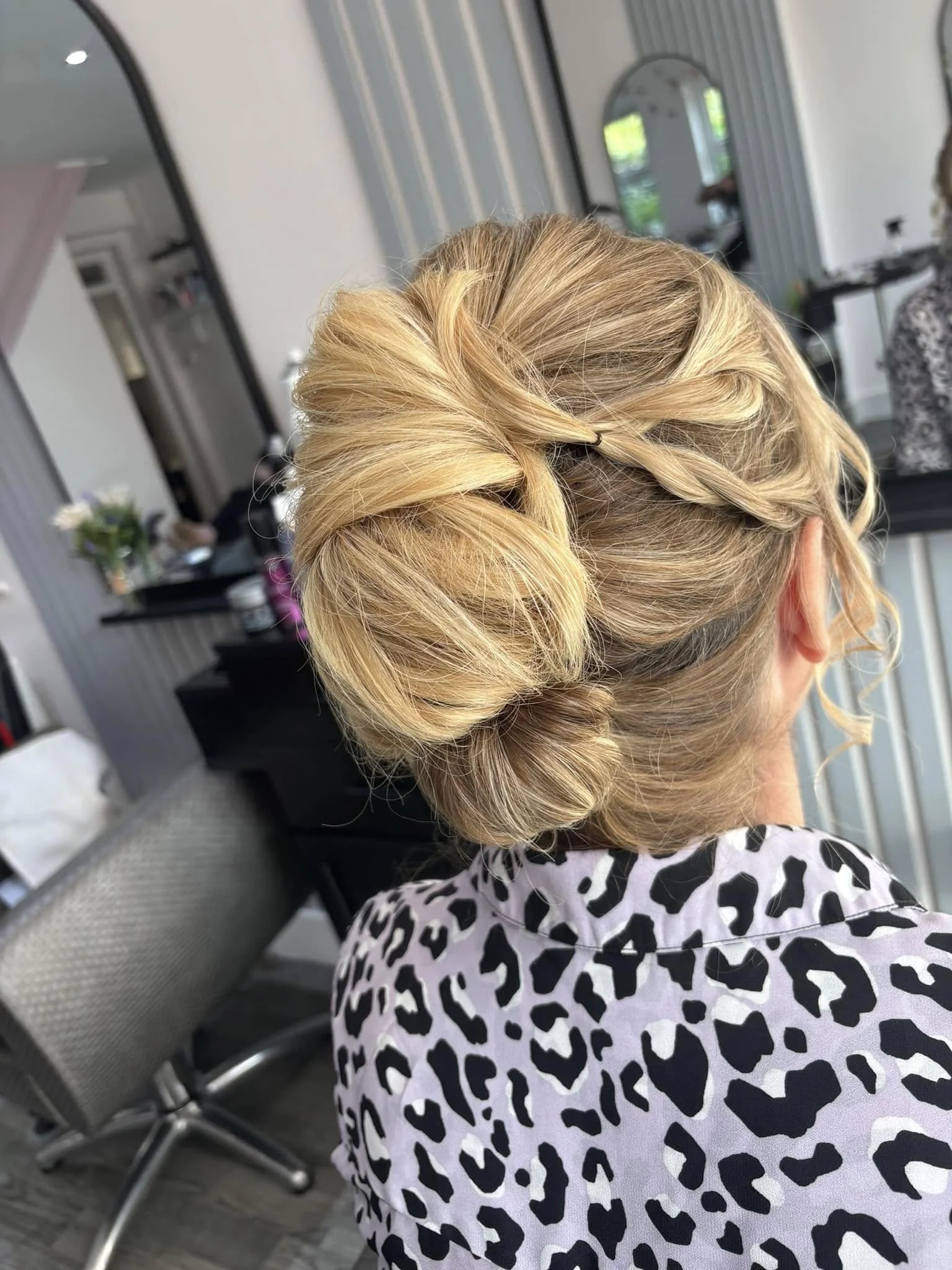 Woman with blonde hair styled in an elegant updo, wearing a black and white animal print top, seated in a salon or salon-like setting.