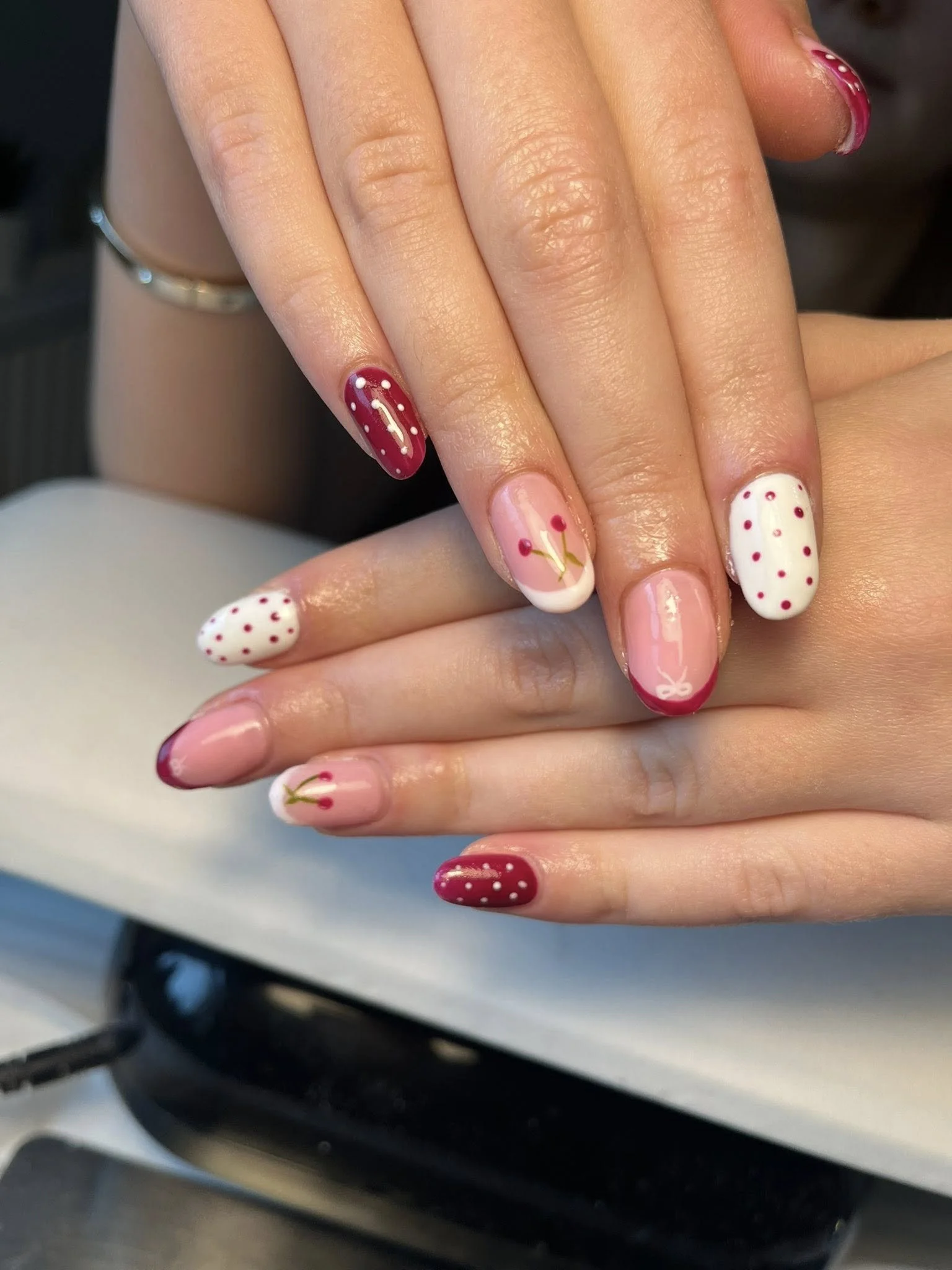 Close-up of two hands with decorated fingernails resting on a surface. The nails have red, white, and pink polish with polka dots and floral designs.