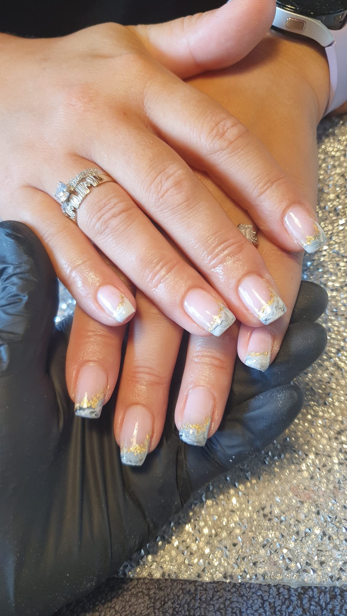 A person's hand with decorated nails resting on another hand. The nails have a French tip design with gold accents and marble-like patterns. A diamond engagement ring and a band with diamonds are on the person's ring finger. The person is wearing a w