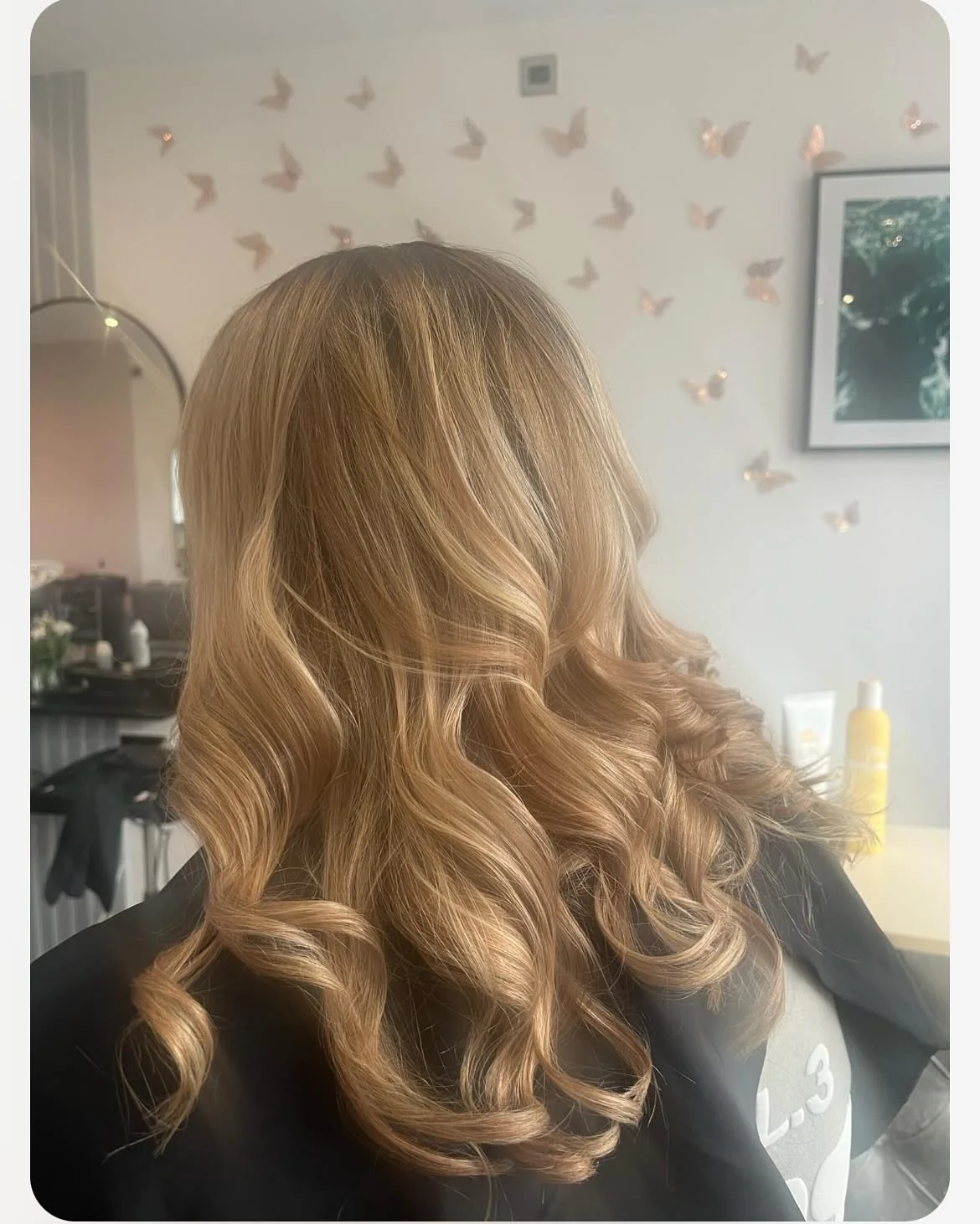 Woman with styled blonde curly hair sitting in a salon chair, with butterfly decorations on the wall, a framed picture, and hair products in the background.
