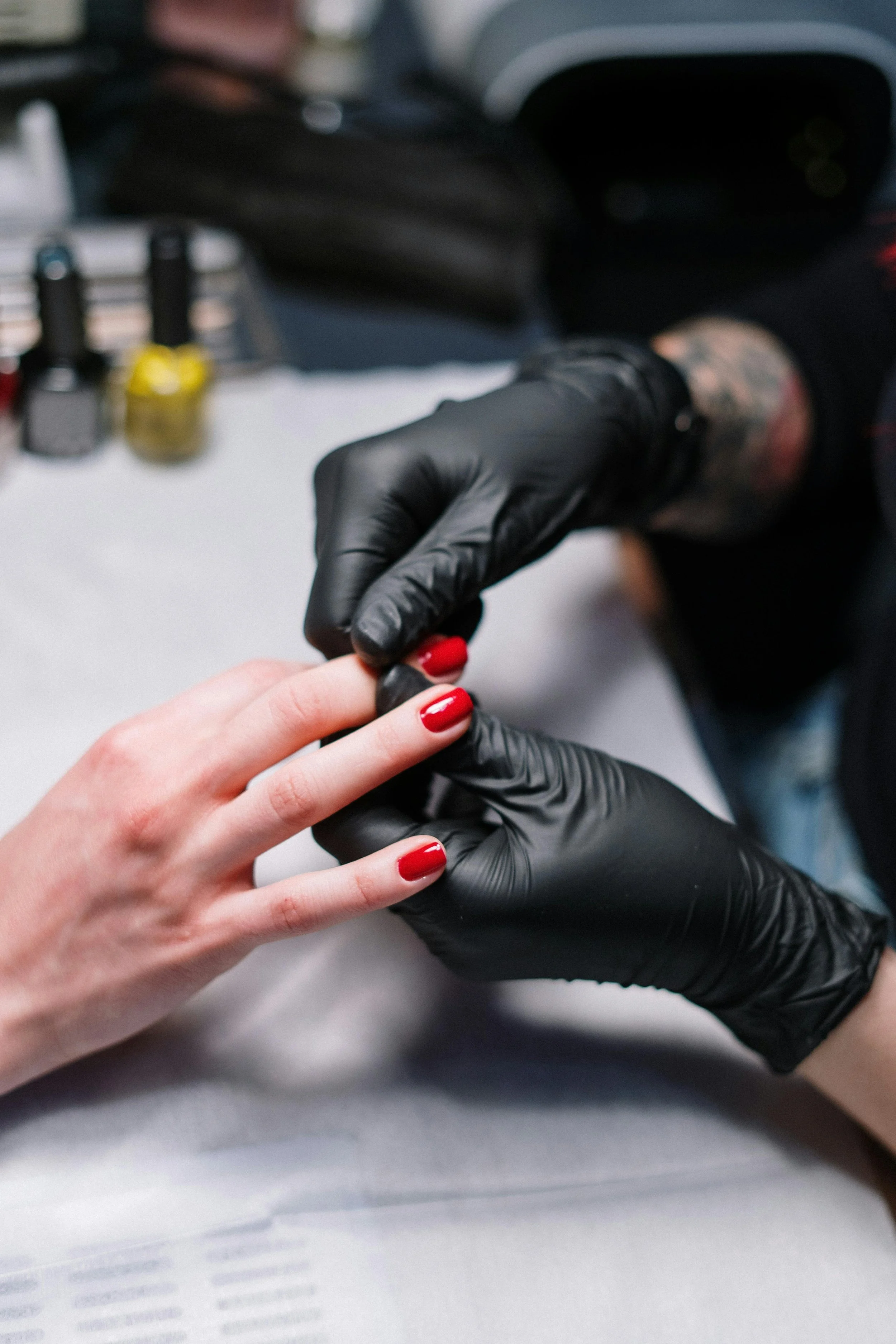 A person getting a manicure with red nail polish at a salon, with the manicurist wearing black gloves, and nail polish bottles in the background.