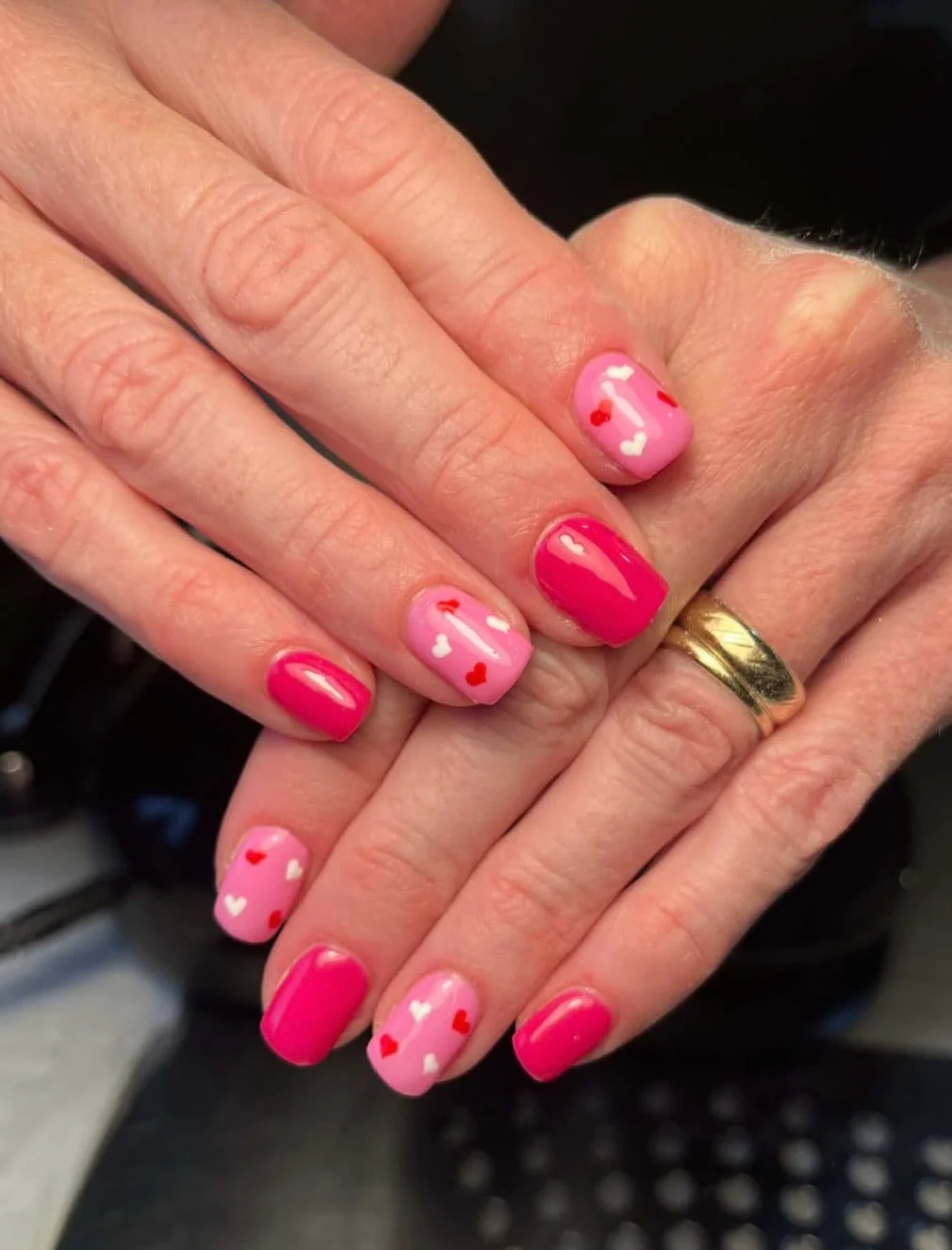 Close-up of hands with manicured nails painted pink with small heart designs, one gold ring, and a black background.