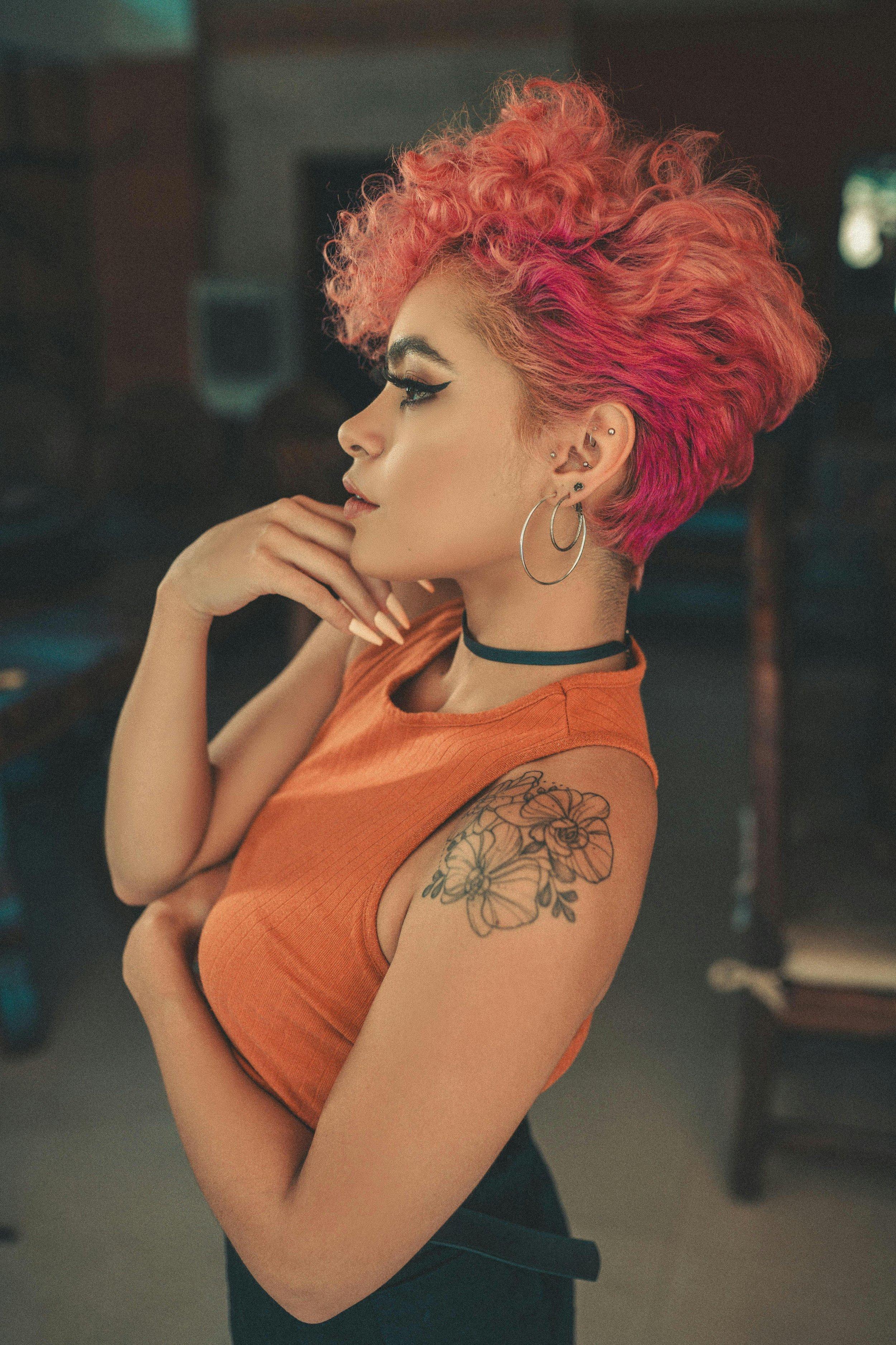 Side profile of a woman with curly, vibrant red and pink hair, wearing hoop earrings, an orange sleeveless top, and a black choker, in an indoor setting.
