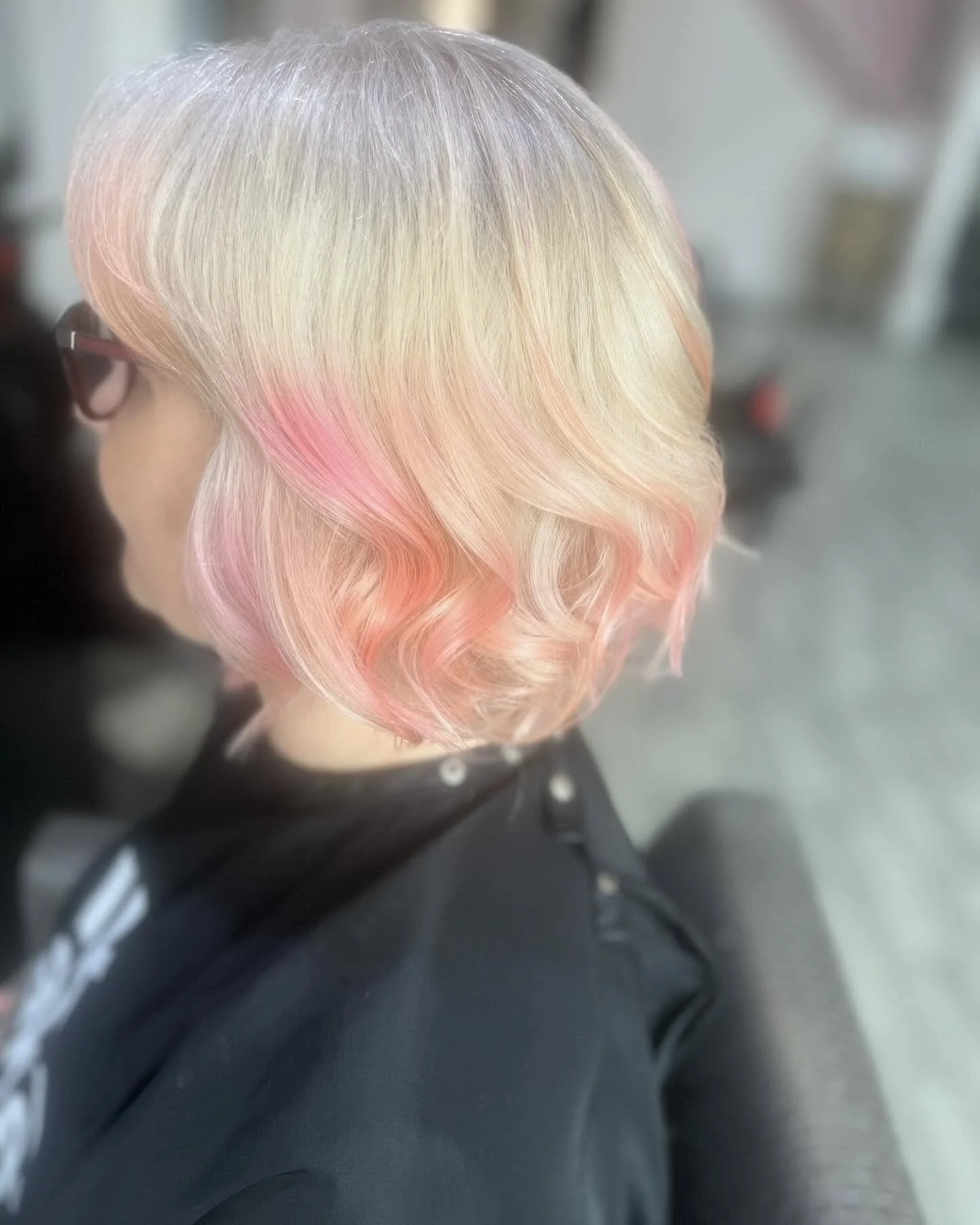 Side view of a person with platinum blonde hair featuring soft pink highlights styled in loose waves, wearing black glasses and a black top.