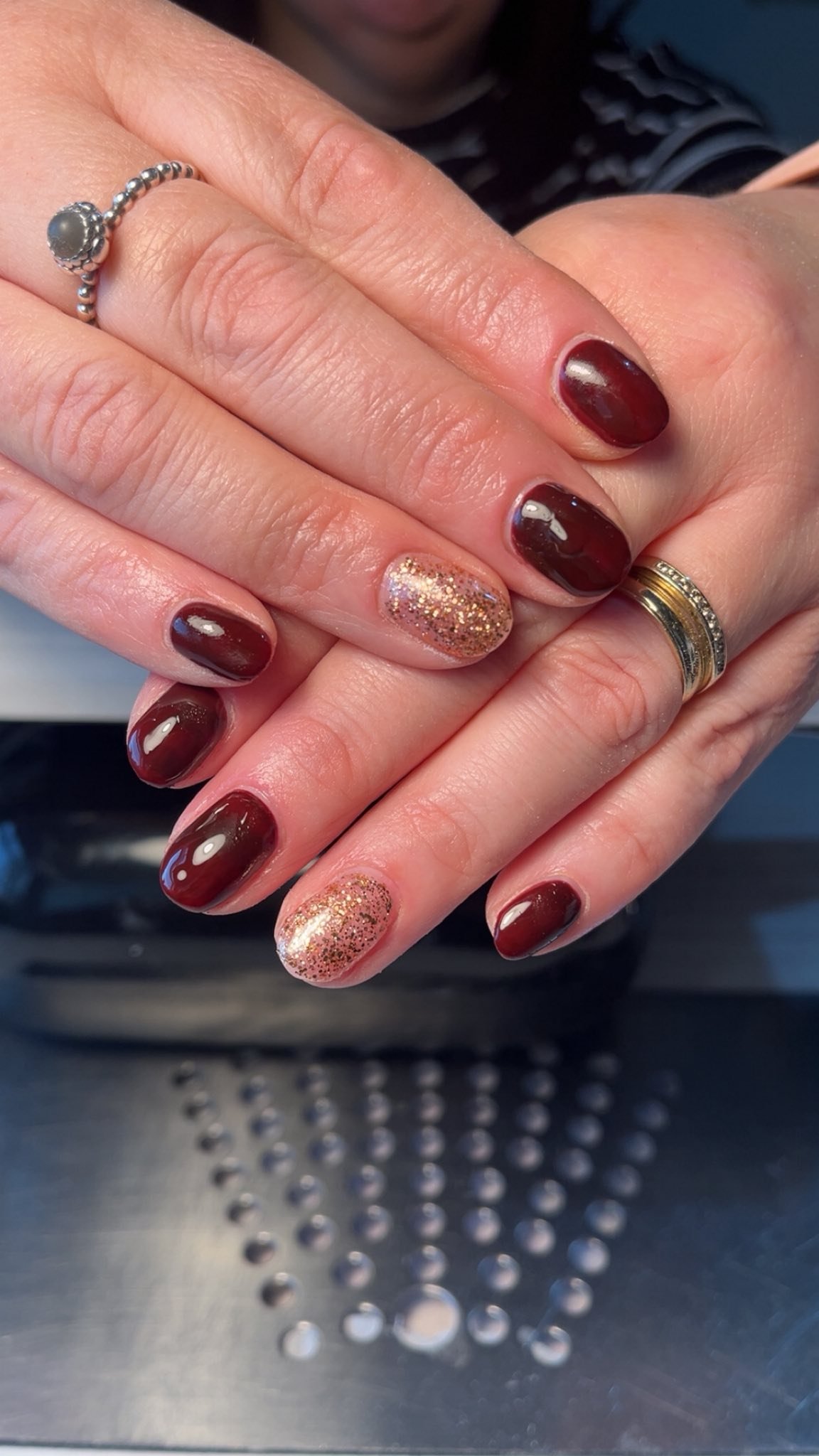Close-up of a person's hands with manicured nails, featuring dark red and glittery gold nail polish. The person is wearing rings, one silver with a gray gemstone and another gold with small gemstones. 