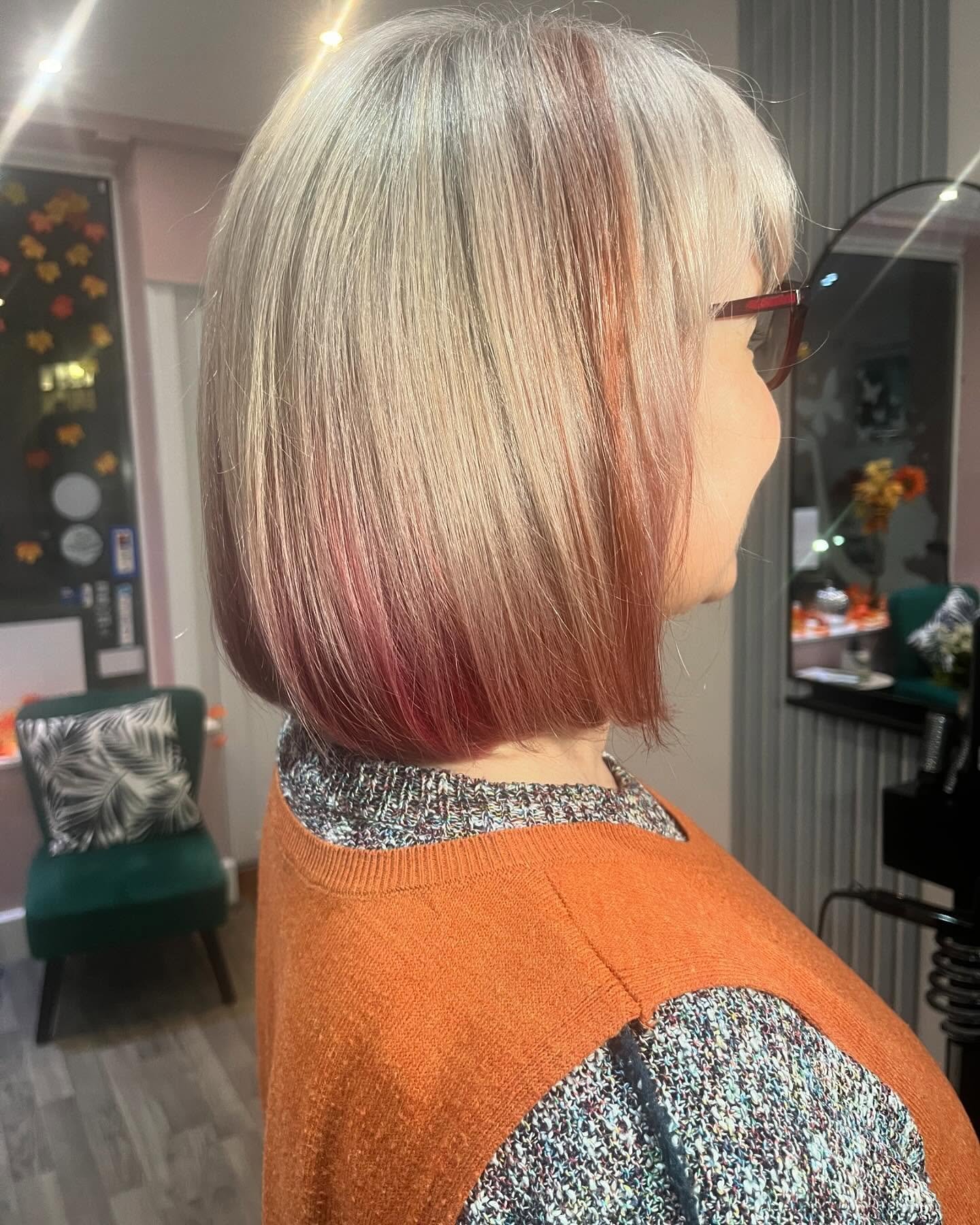 Side view of a woman with shoulder-length blonde hair with pinkish tips, wearing glasses and an orange sweater.