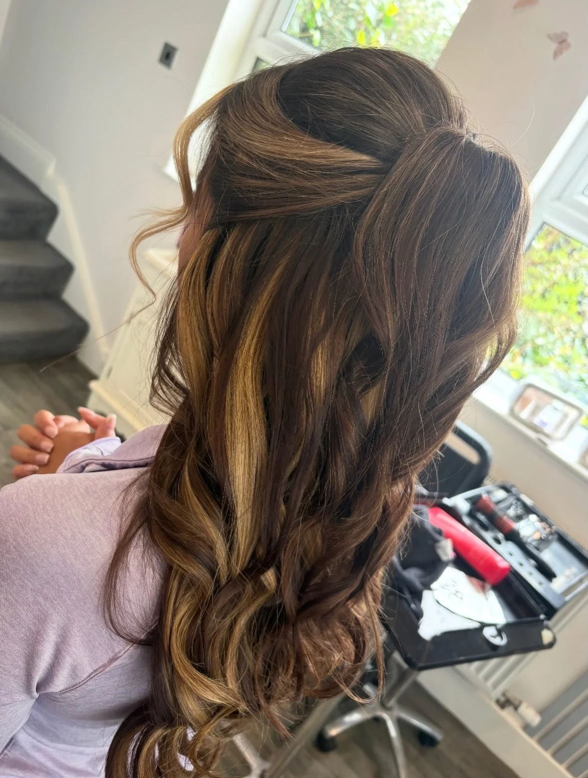 A woman with long, wavy, brown hair with blonde highlights, sitting indoors in front of a window, with curling iron tools and a case on a table nearby.