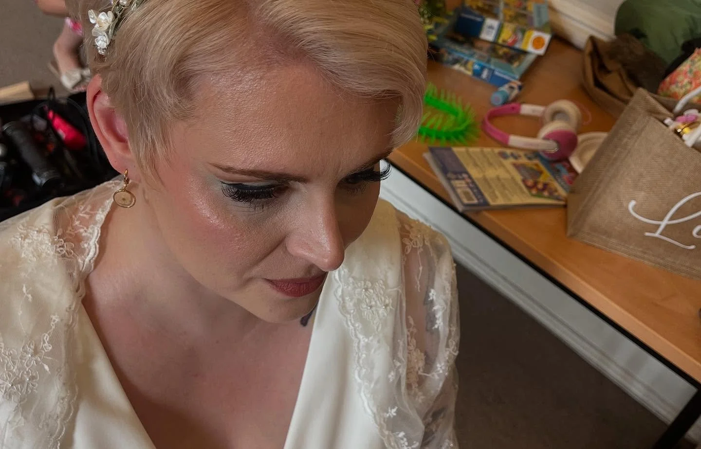 A woman with short blonde hair and earrings, dressed in a cream-colored satin dress with lace details, looking down. Lisa B Hair & Beauty Salon, Carlisle, Cumbria UK.
