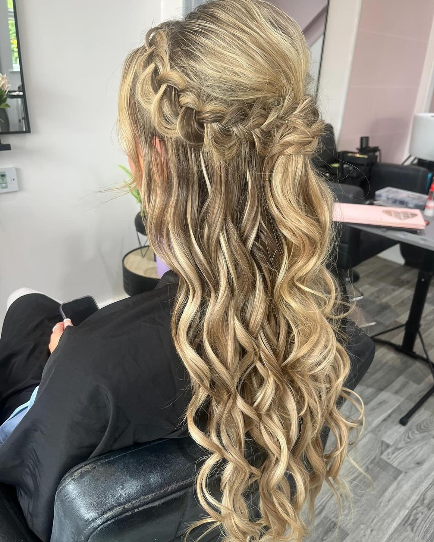A woman with long, blonde, curly hair styled with braided crown and loose waves, sitting in a salon chair.