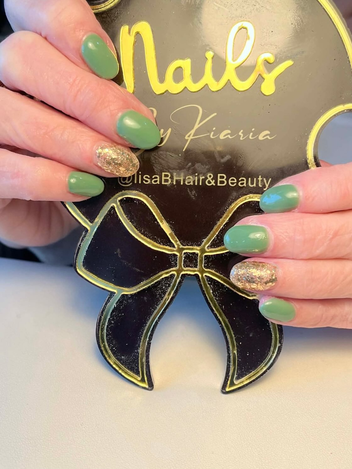 Person holding a decorative plaque with the text 'Nails by Kiaria' and Instagram handle '@LisaBHair&Beauty', with green and glittery gold painted nails.