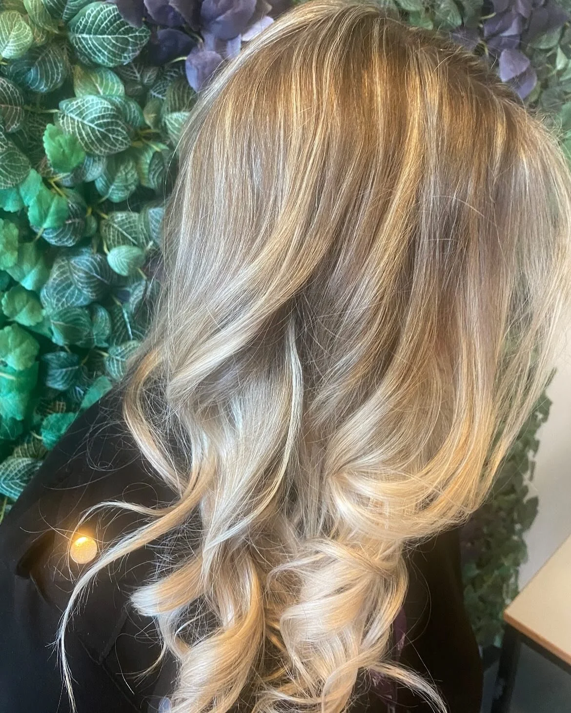 Blonde hair styled in loose curls against a green leafy background.