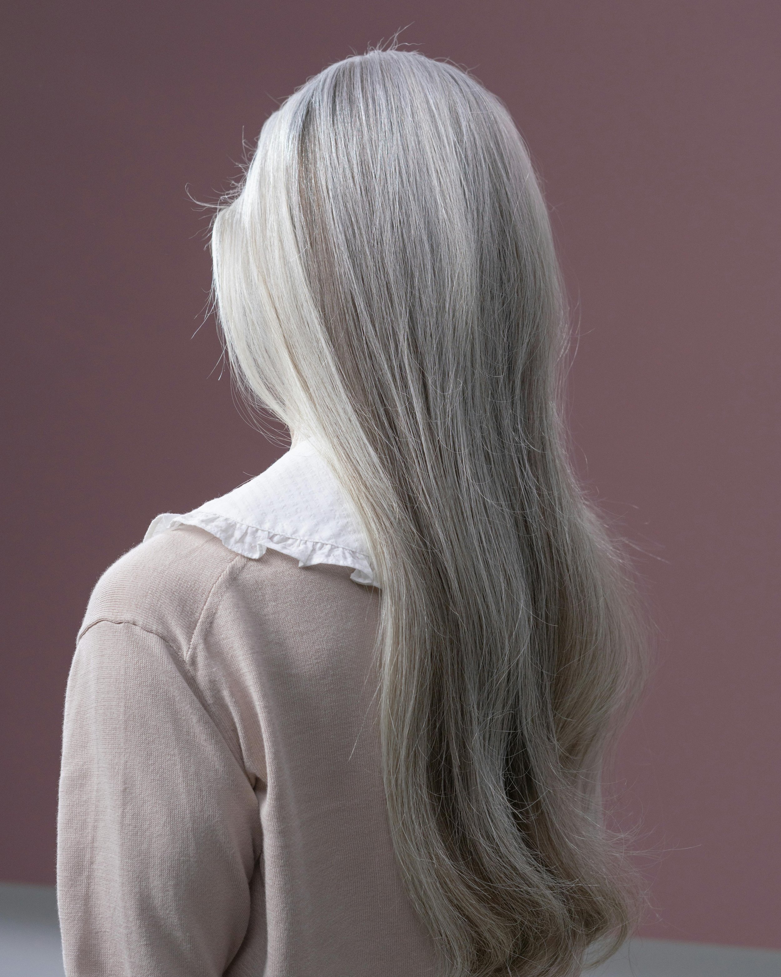 Back view of a person with long, straight, silver hair wearing a light beige sweater with a white ruffled collar.