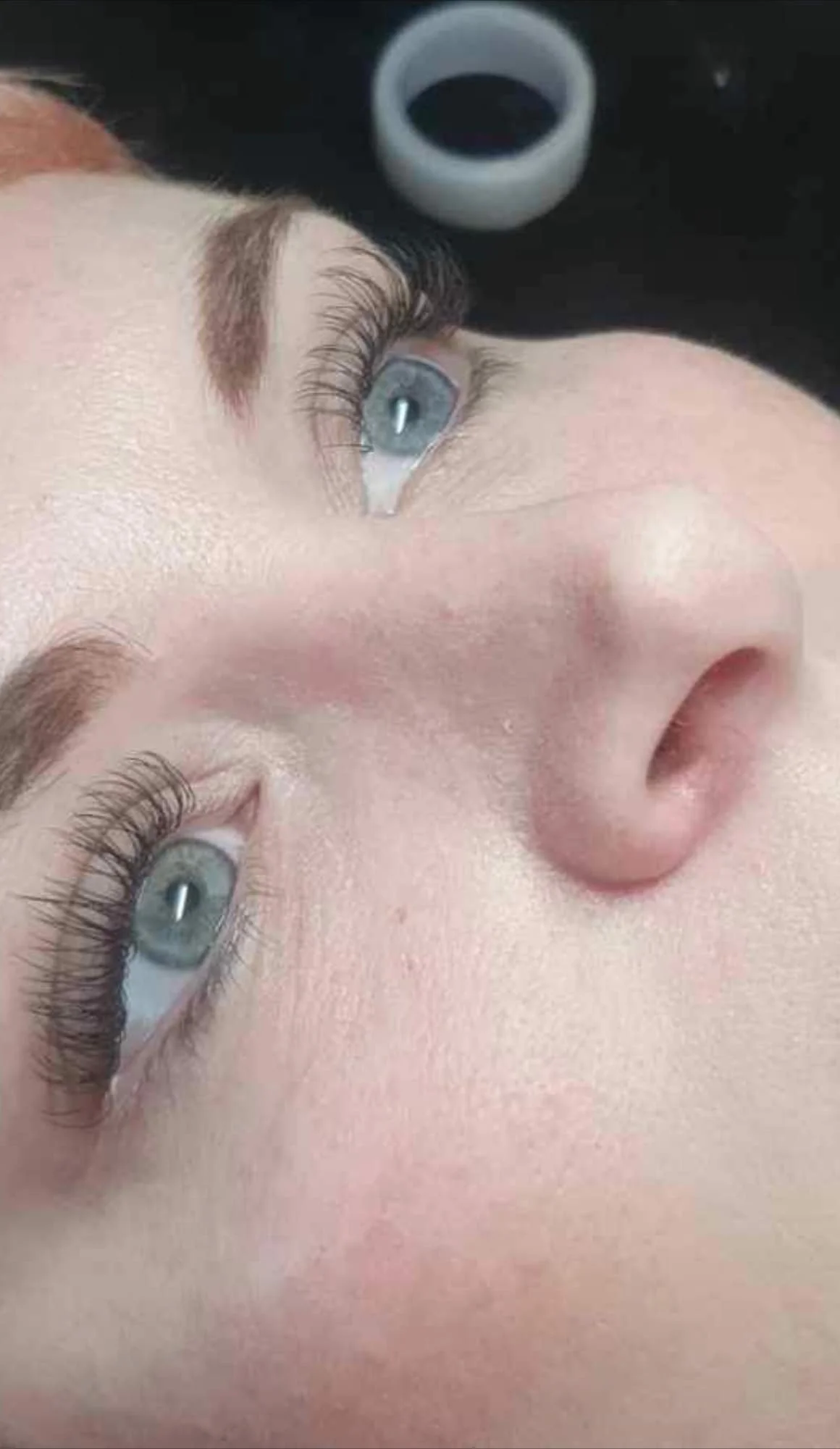 Close-up of a person with blue eyes, long eyelashes, fair skin, and red eyebrows, lying down with a black background and a small round white object nearby.