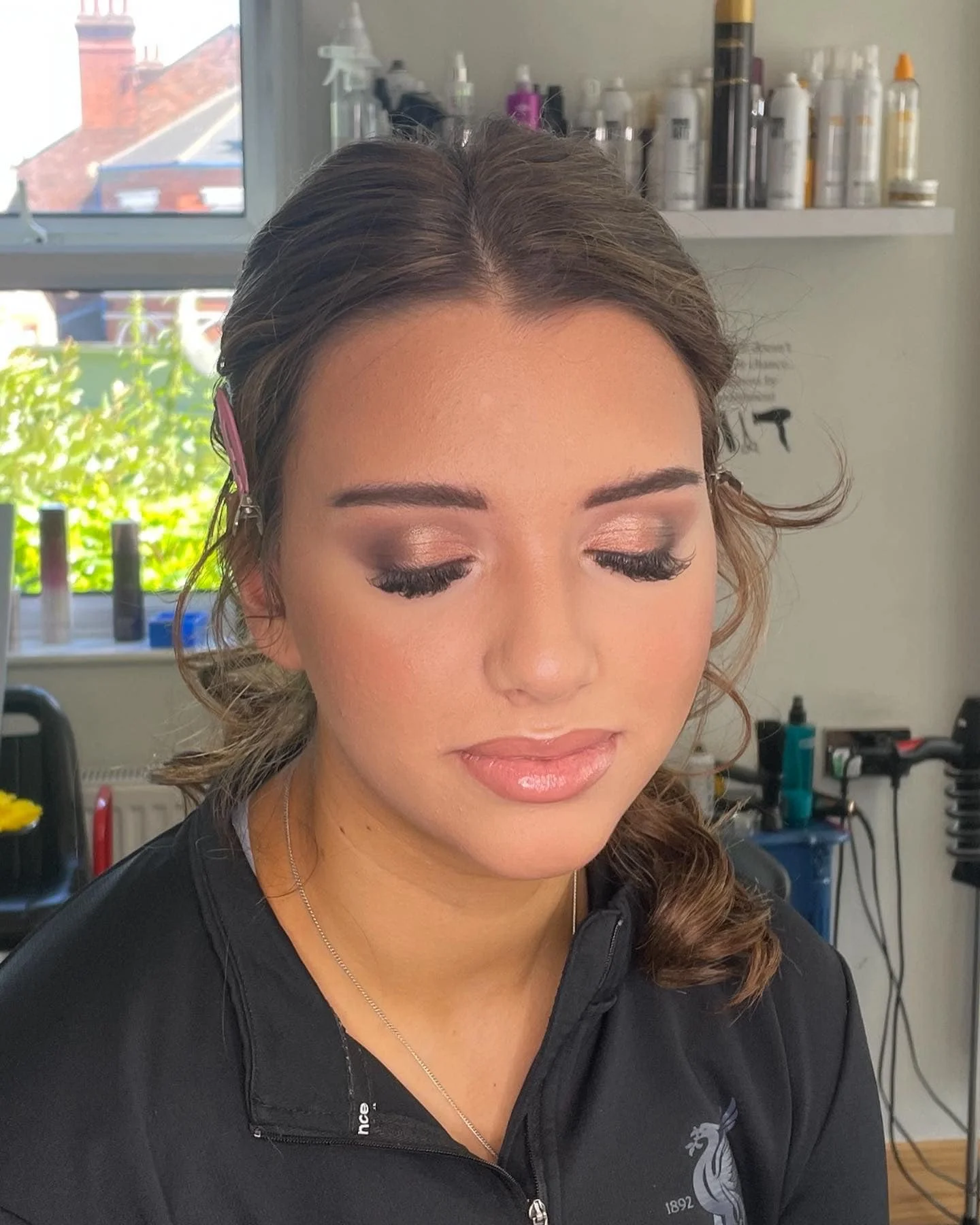 A woman with light skin and long curly brown hair, wearing a black jacket with a lion emblem, sitting in a room with a large window behind her. at Lisa B Hair & Beauty Salon, Carlisle, Cumbria UK.