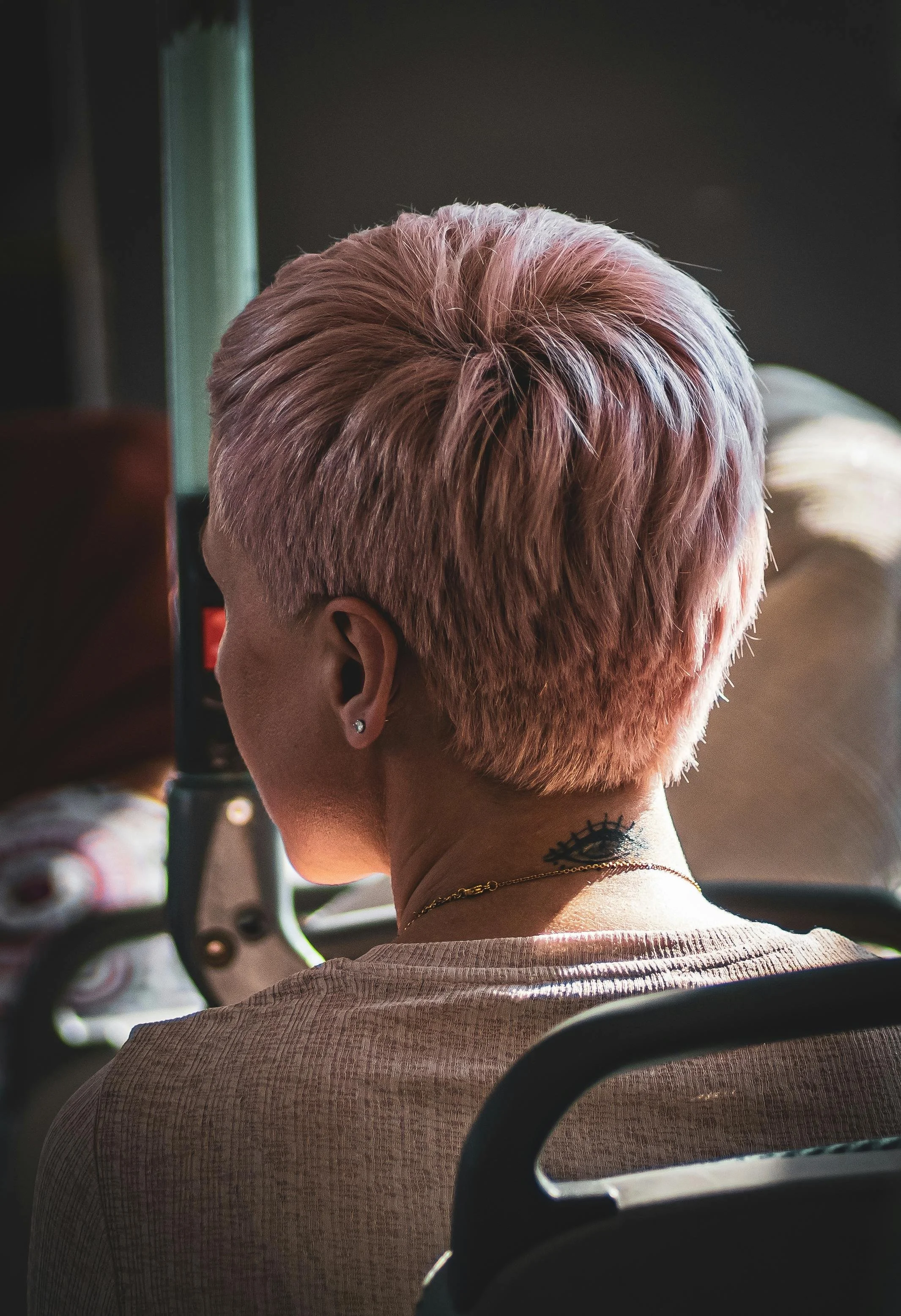 Close-up of a person with short, pastel pink hair, sitting in a vehicle, wearing a beige shirt, a necklace, an earring, and a tattoo on the neck.