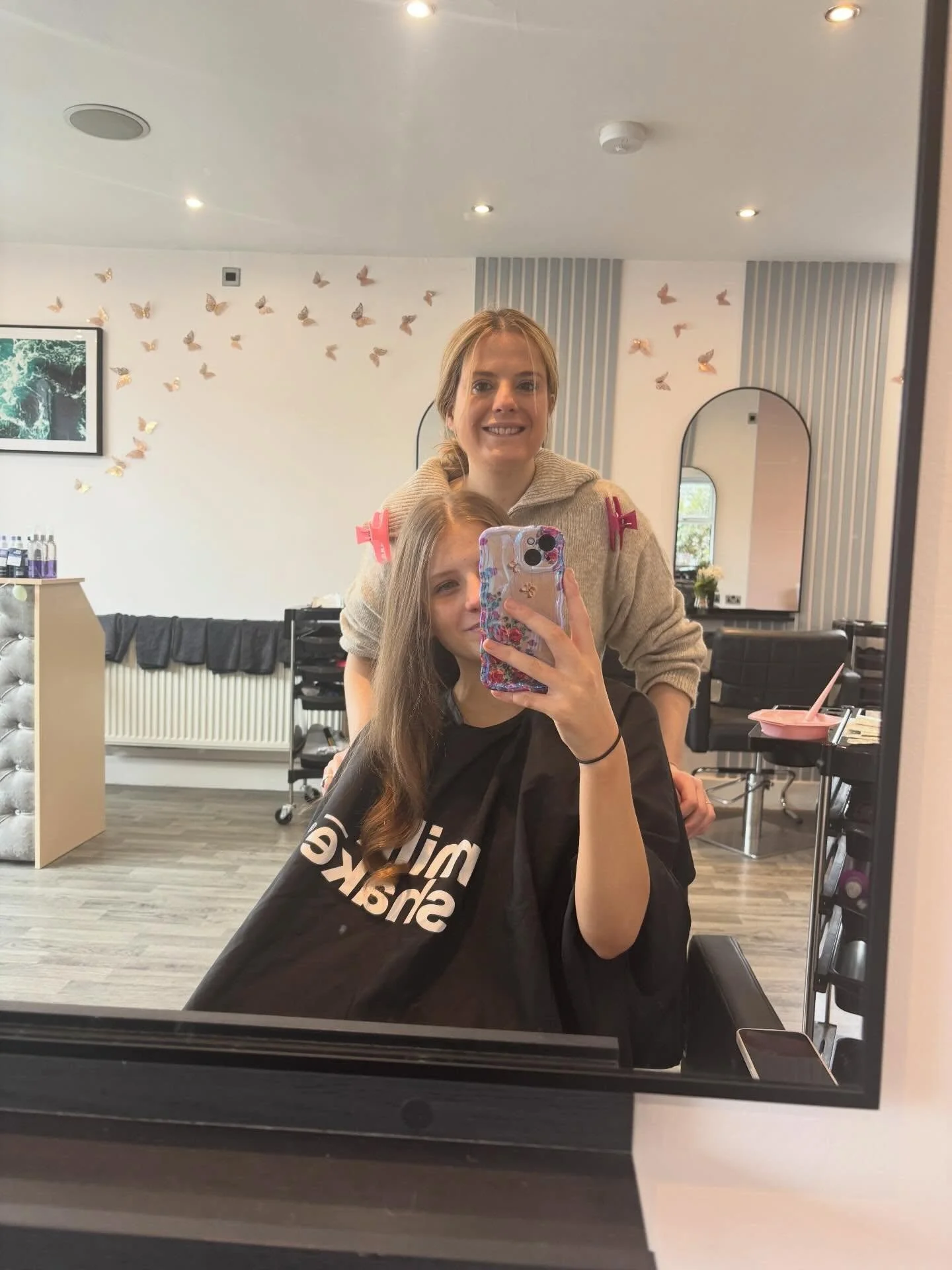 A woman taking a selfie in a hair salon mirror with a young girl sitting in front of her. The salon has butterfly decorations on the wall, a painting, and salon chairs.
