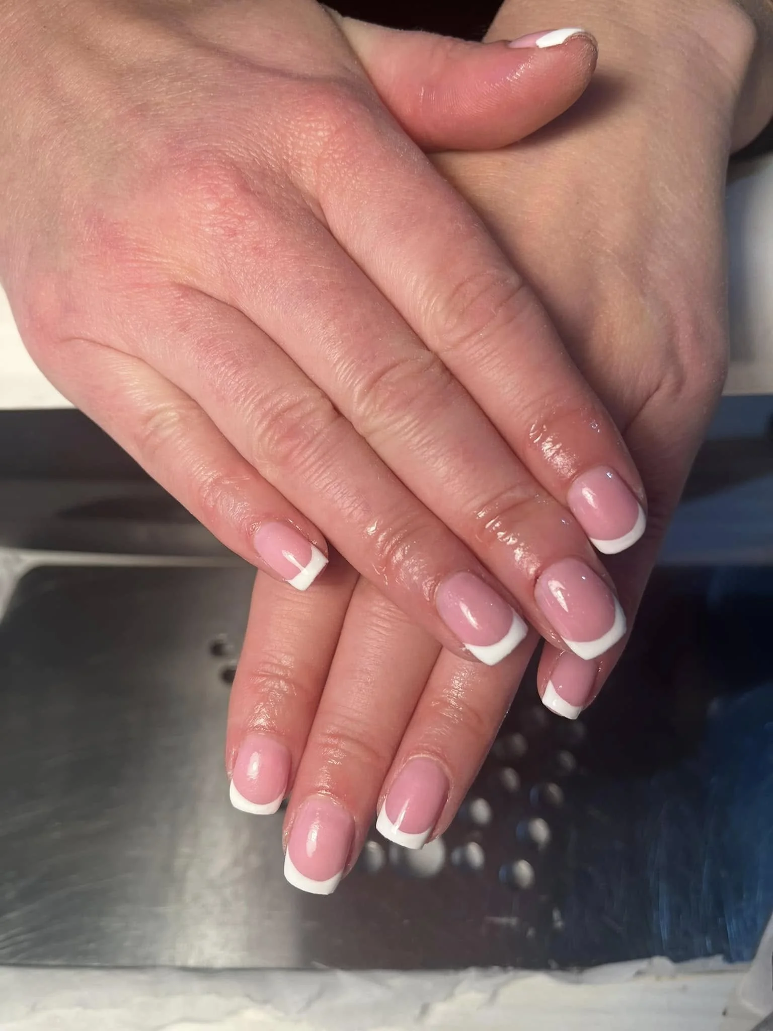 Close-up of hands with neatly manicured nails featuring a French tip design, resting on a metallic surface. Award winning Lisa B Hair and Beauty Salon in Carlisle, Cumbria UK.