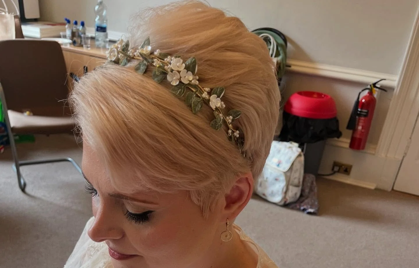 A woman with blonde hair wearing a floral headband and earrings, in an indoor setting with a desk and fire safety equipment in the background.