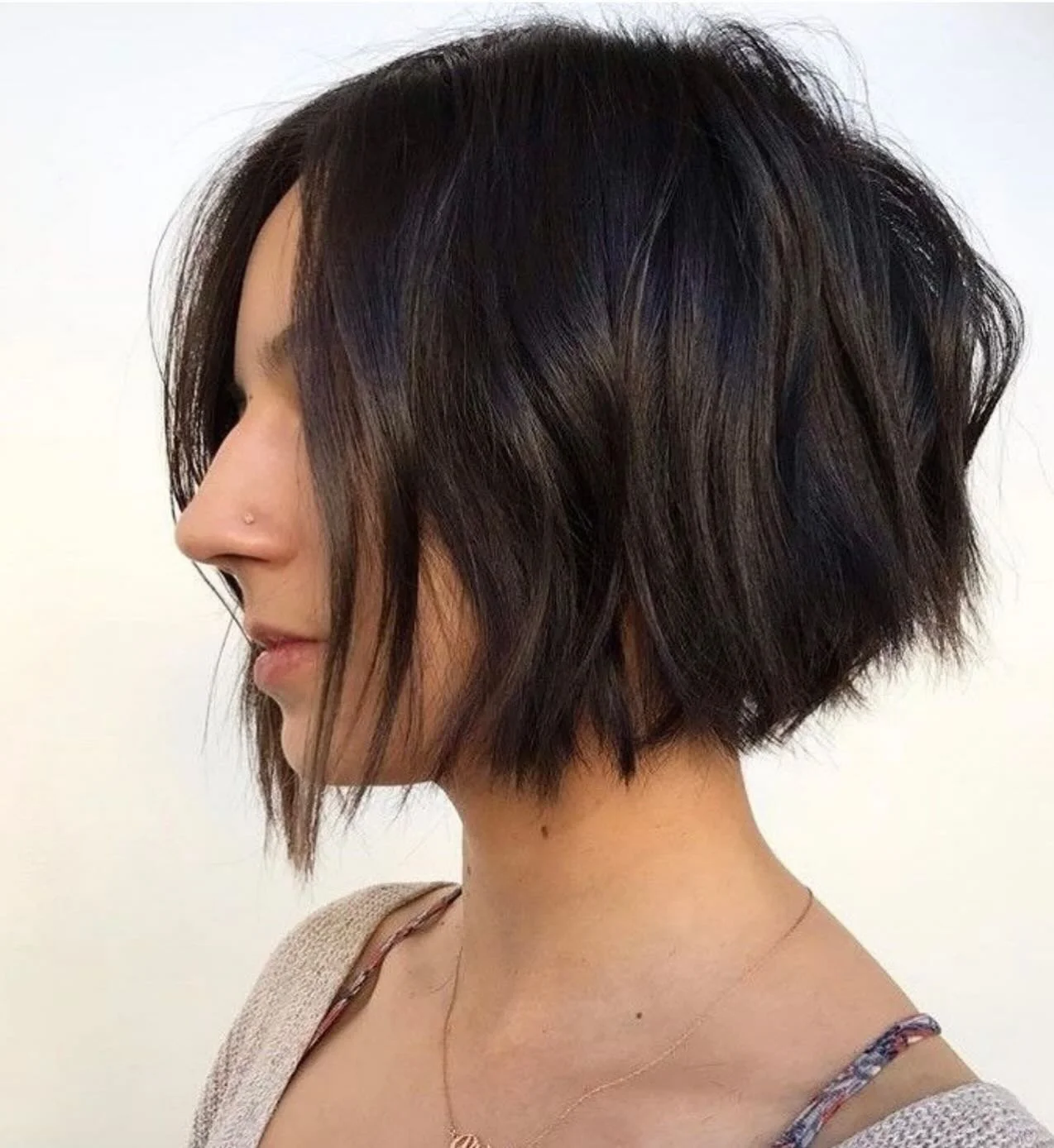 Side profile of a woman with a short, layered, dark brown bob haircut with some lighter highlights, wearing a beige top and a delicate necklace.