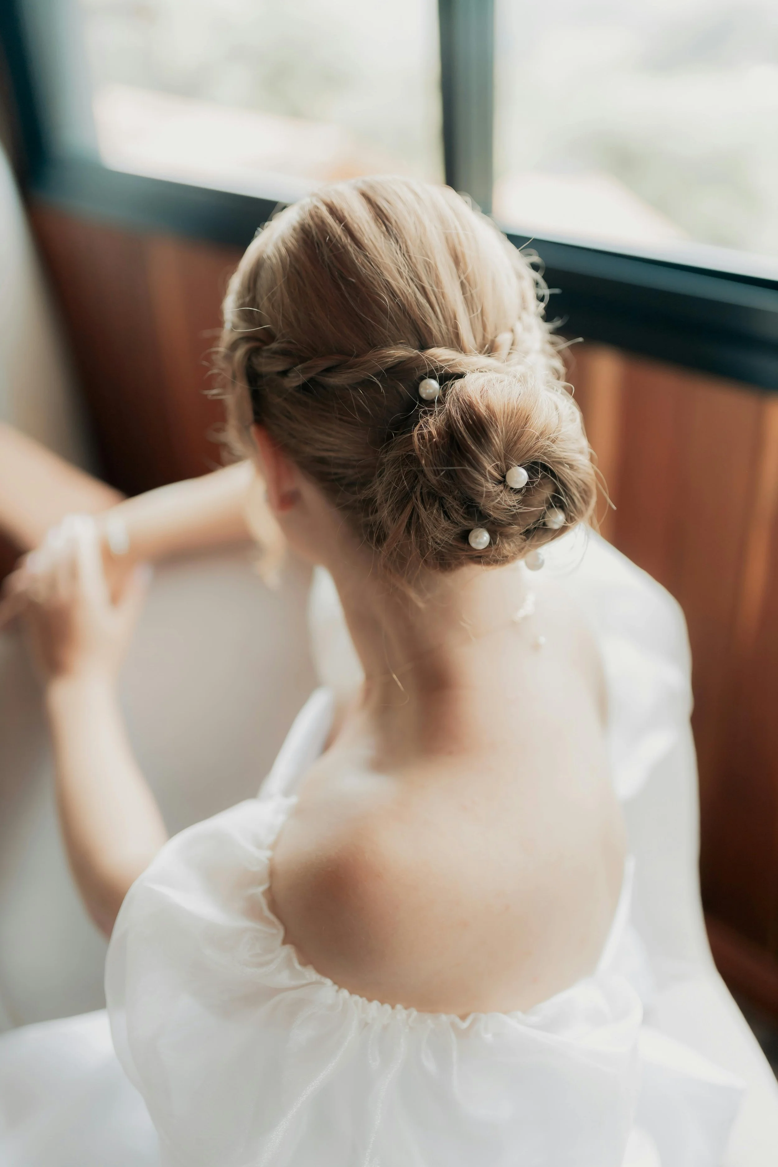 A woman with blonde hair styled in an elegant updo adorned with pearl hairpins, wearing a white dress with puffed sleeves, sitting by a window with natural light.