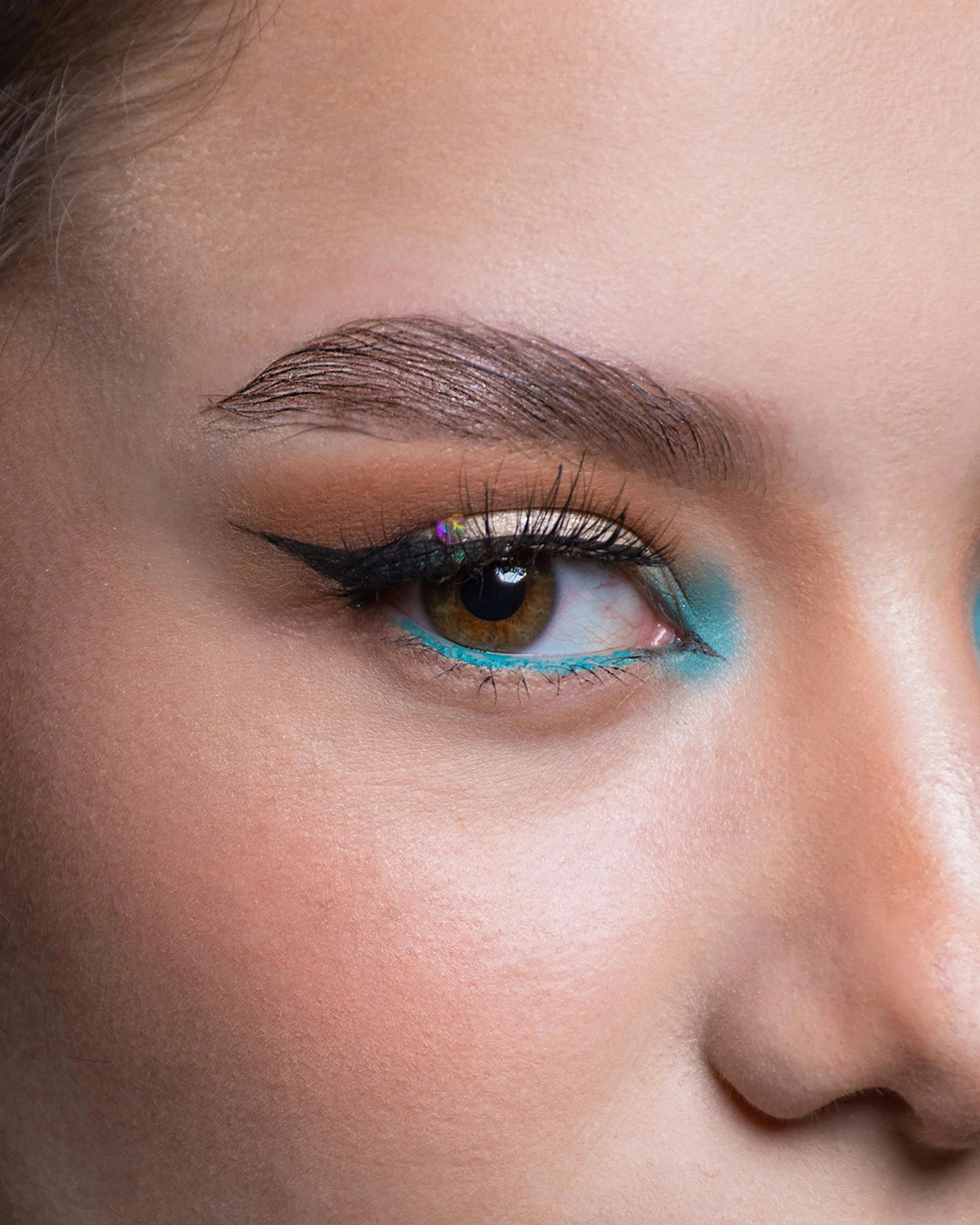 Close-up of a person’s eye with makeup and a brown eyebrow, with a blue eyeliner and small rhinestones near the eye.