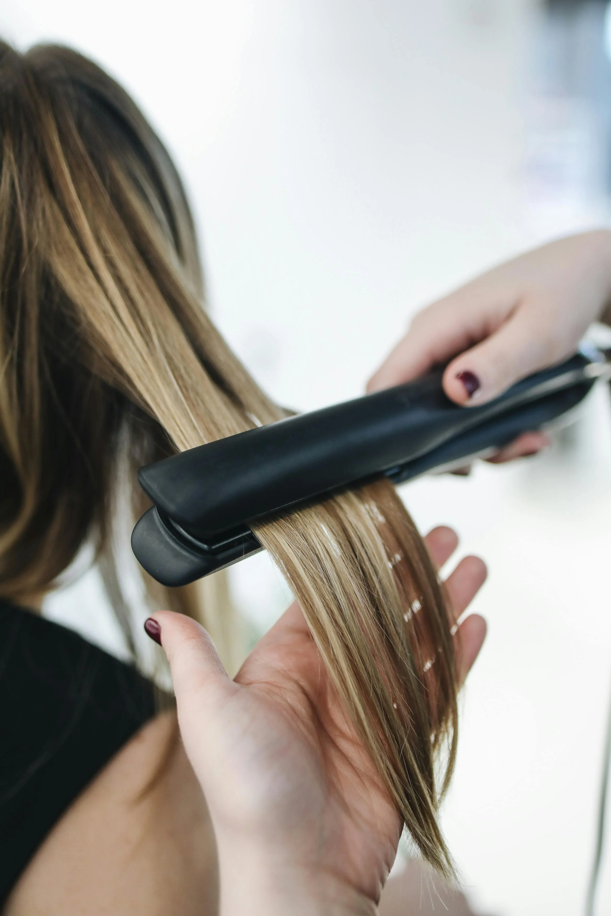 A person with long, blonde hair getting a haircut at a salon, with a stylist using flat iron styling the hair.