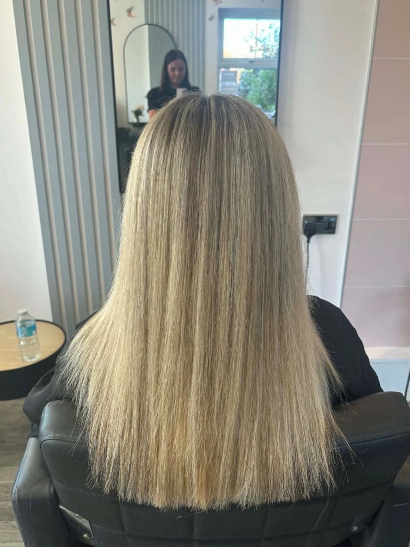 Back view of a woman with long, straight blonde hair sitting in a salon chair, facing a mirror, with a window in the background.