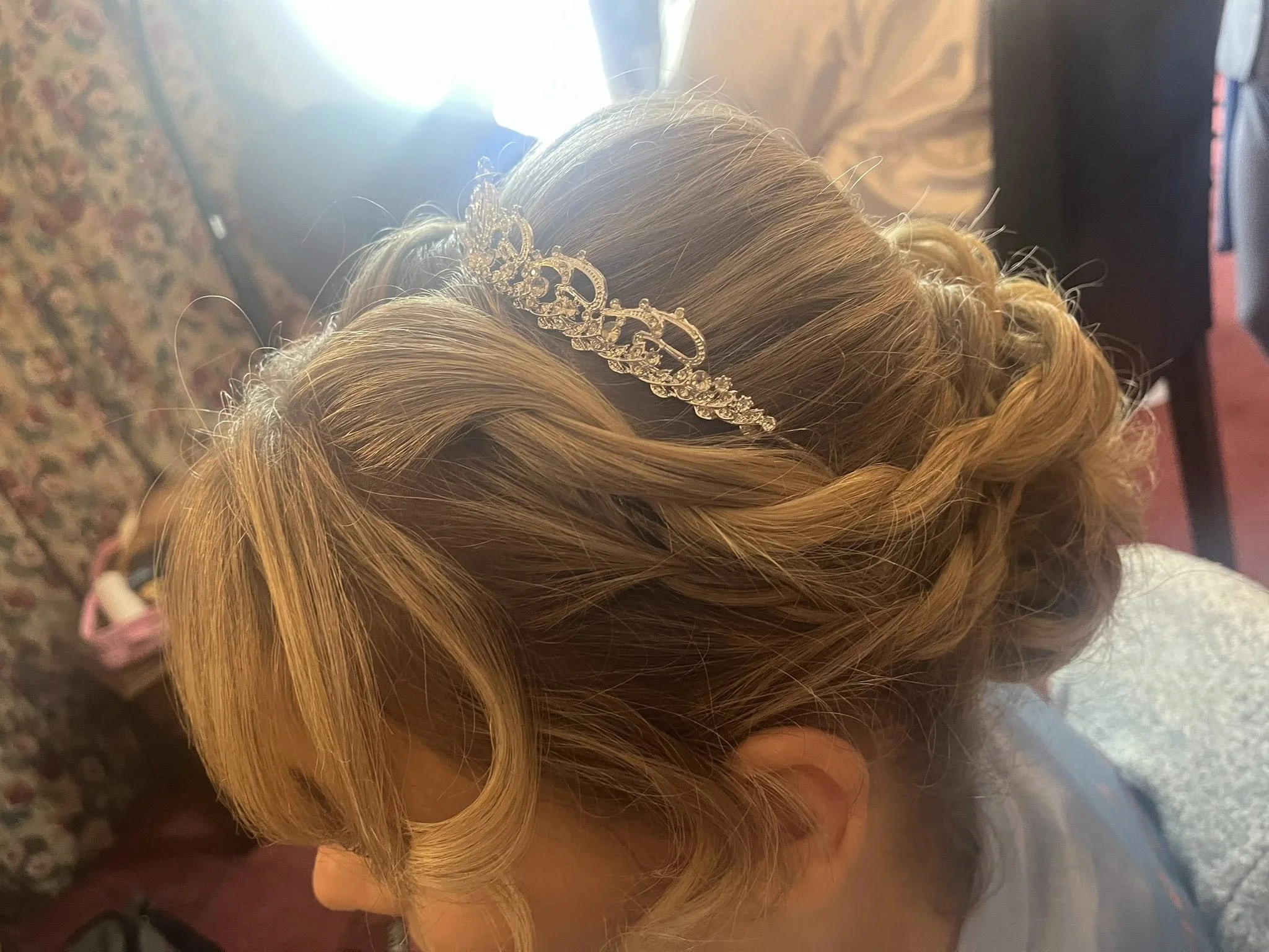 Close-up of a person's styled blonde hair with loose curls, featuring a decorative silver hair accessory with intricate details, against a background including a floral-patterned fabric.