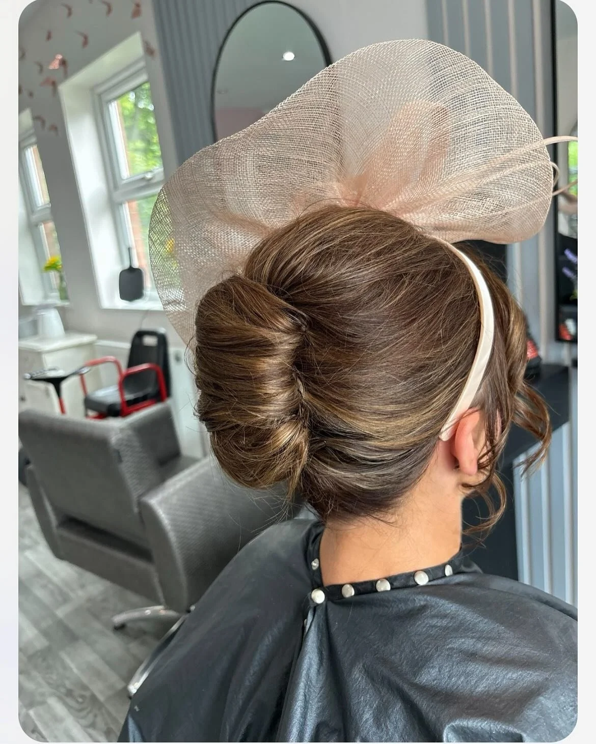 A woman with neatly styled brown hair in an elegant bun, wearing a large light pink fascinator with mesh material, and a headband, sitting in a salon chair with a black cape with white pearls around her neck.