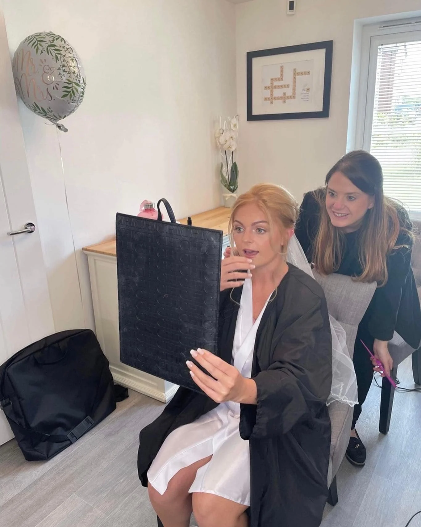 A woman dressed in a white gown and black robe sits at a desk, looking at a computer screen with a surprised expression at Lisa B Hair & Beauty Salon, Carlisle, Cumbria UK.