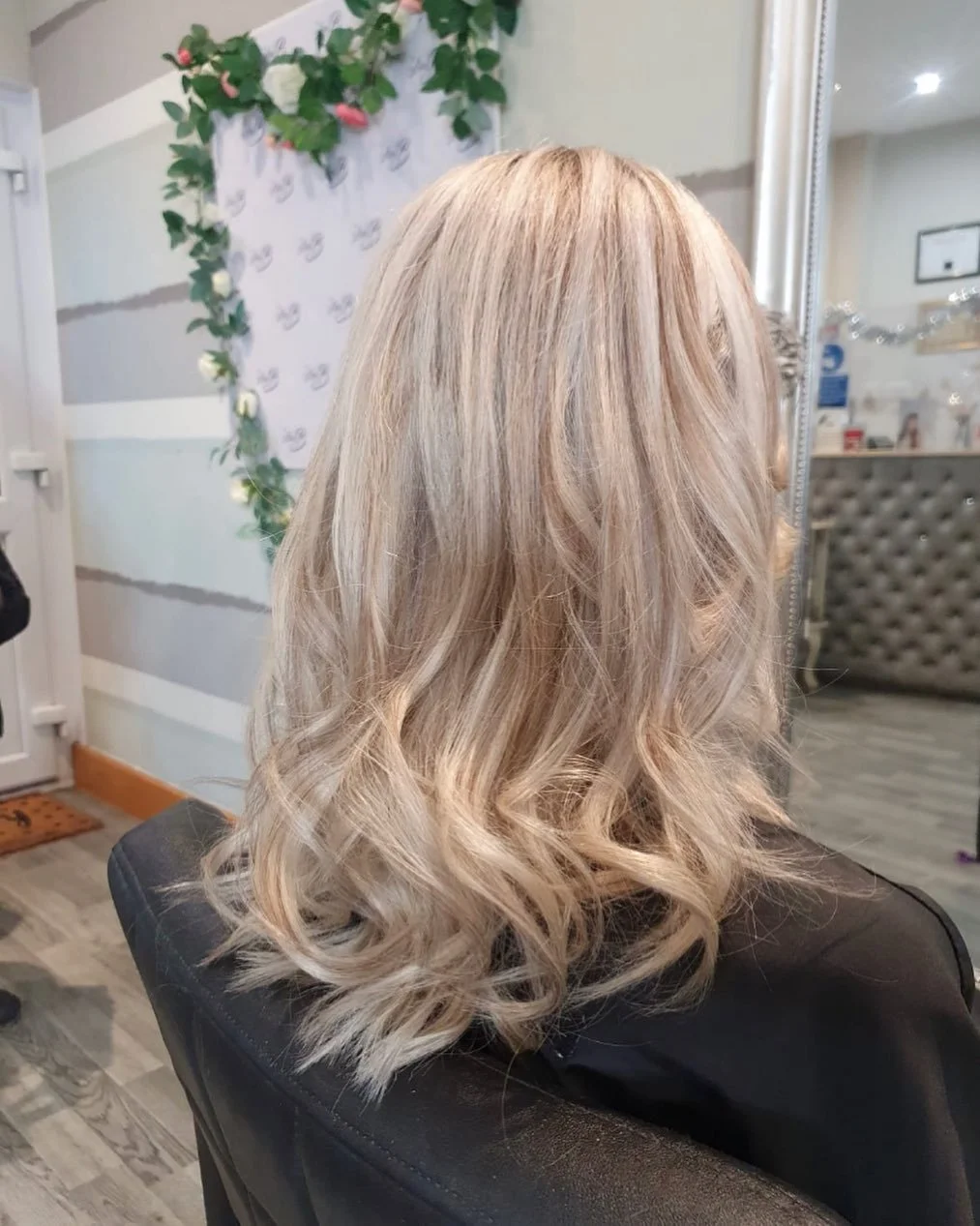 Back of a woman with shoulder-length, wavy, blonde hair sitting in a salon chair in front of a mirror, with decorative greenery on the wall in the background.