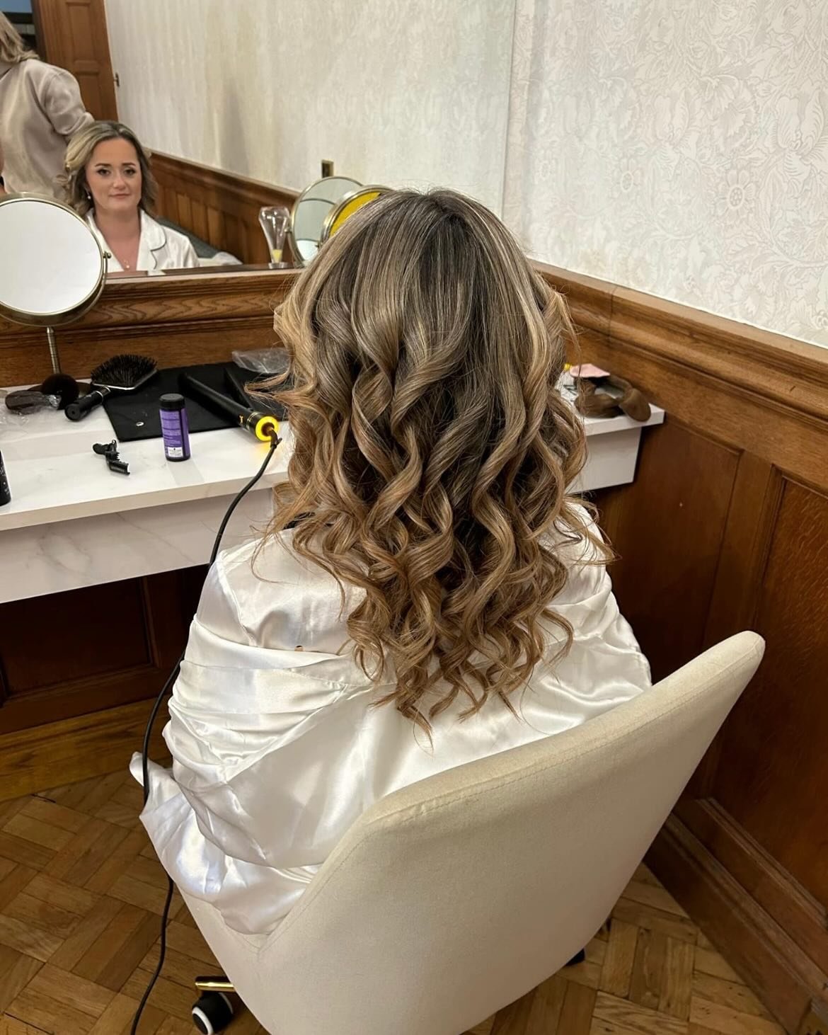 A woman with styled, loose curls hair sitting in a salon chair in front of a mirror, with her back visible. The salon mirror reflects her face and part of the room, with various hairstyling tools on the table.
